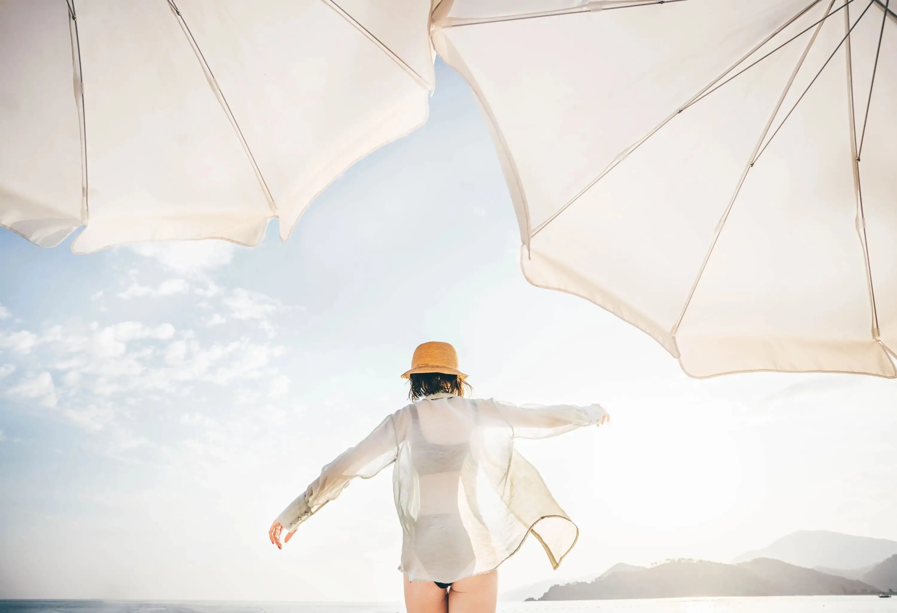A woman in a bathing suit, straw hat, and see-through long-sleeve stands under the sun beside two white umbrellas.