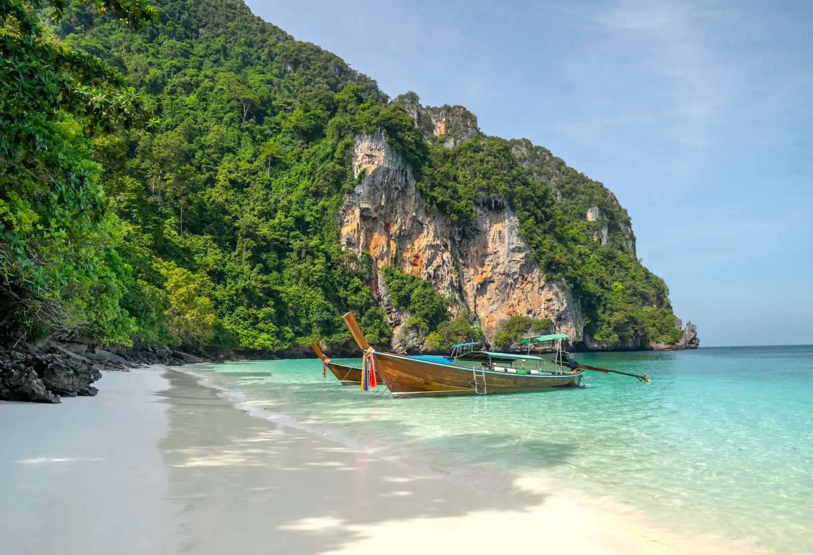 Two long-tail boats anchored on a white beach along a steep and rocky coastline covered in dense vegetation.