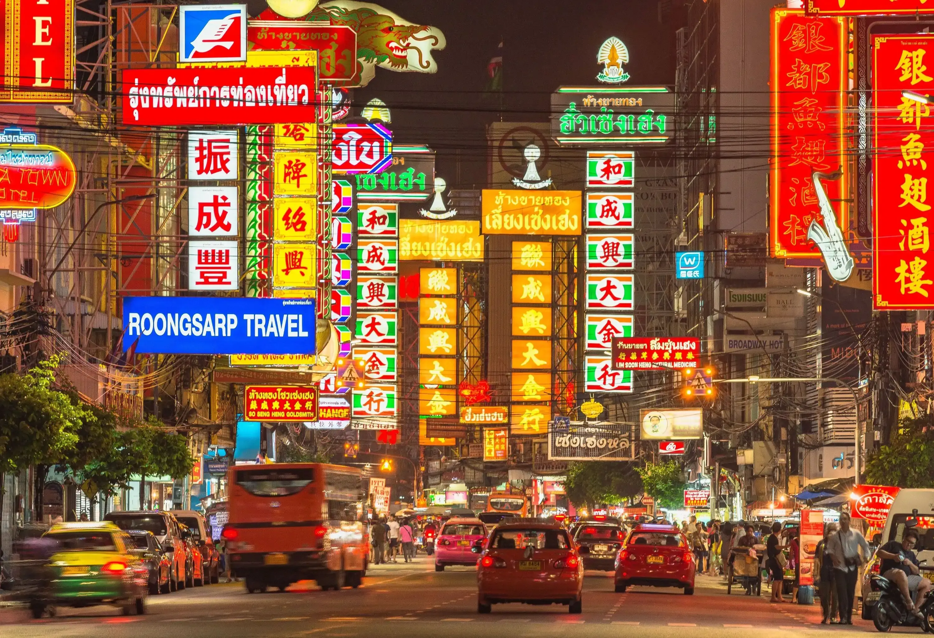 A busy highway with cars passing and pedestrians walking on the side under the bright signages at night.