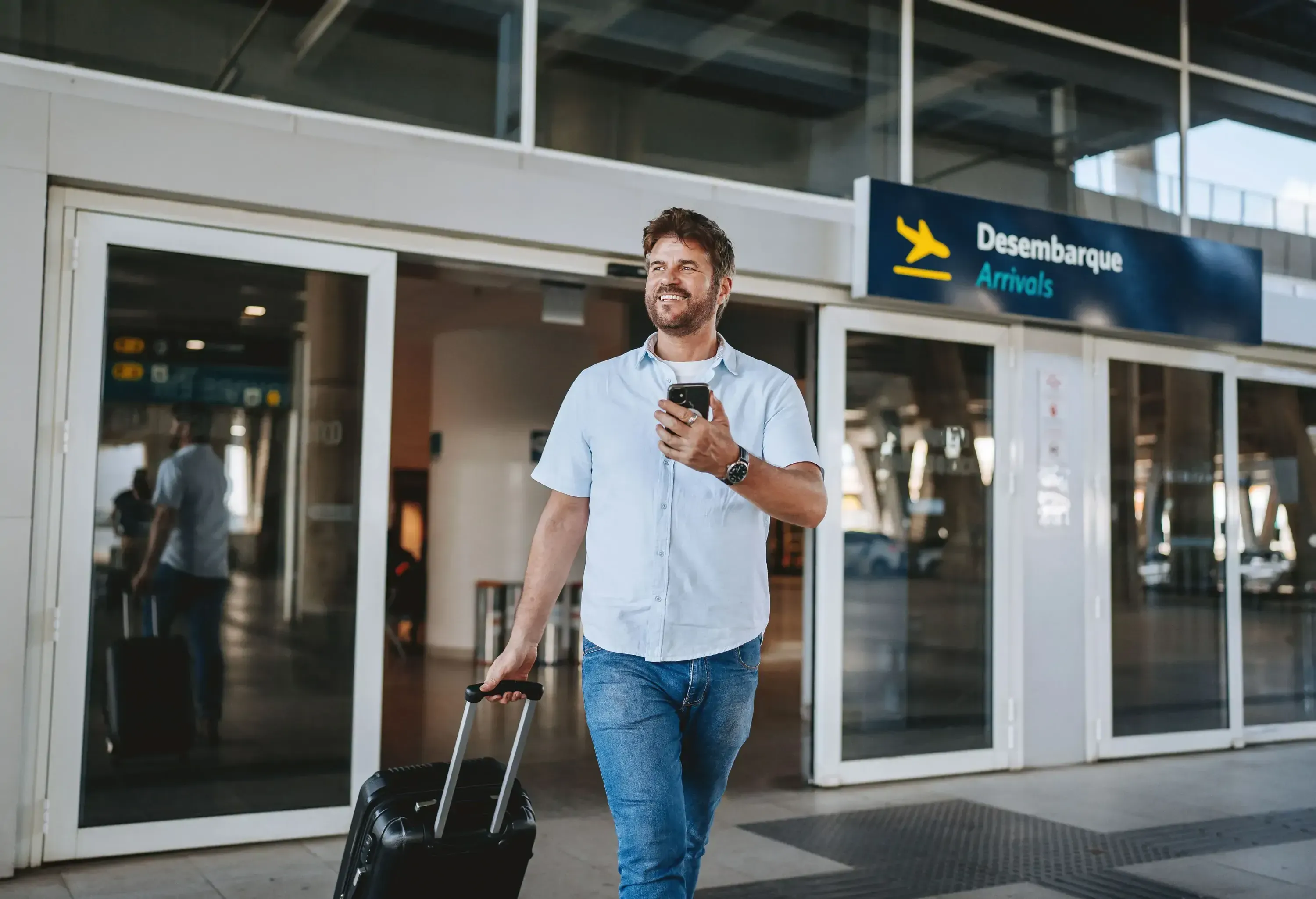 Man smiling and walking out of airport arrivals, pulling a suitcase and holding a phone.