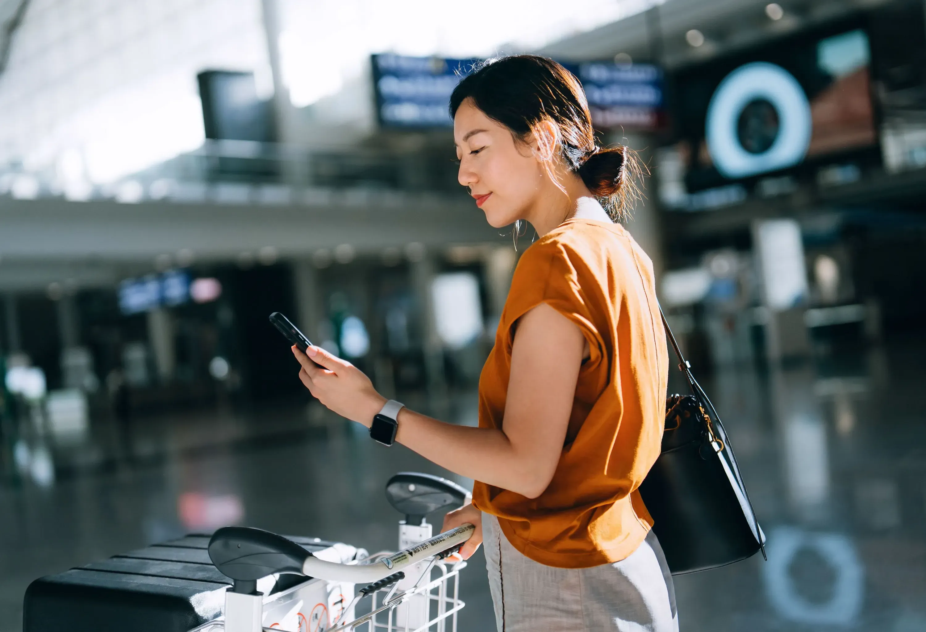 Young Asian woman using smartphone and pushing a luggage trolley with suitcases at airport terminal.