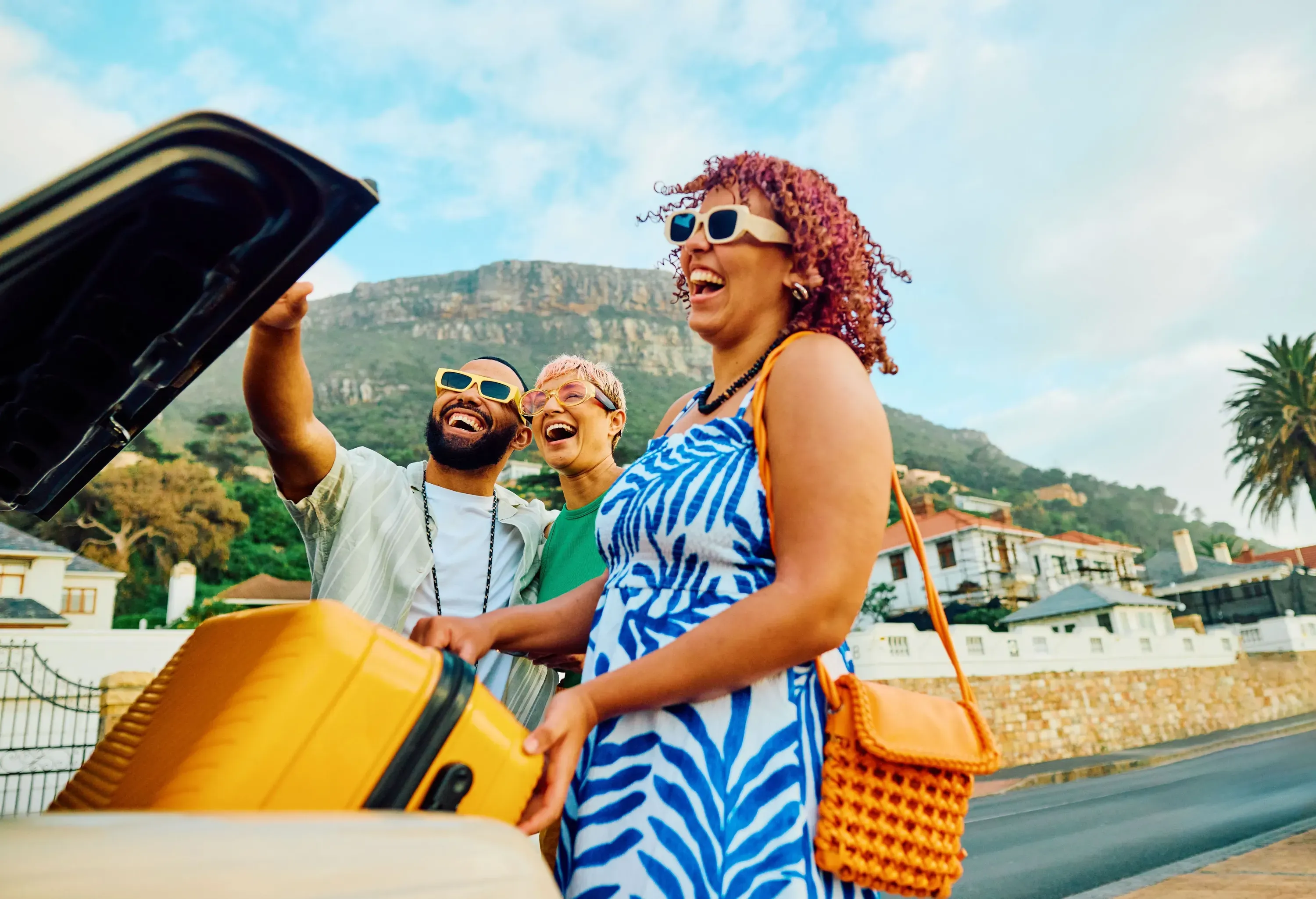 Three happy people laughing while packing luggage into a car trunk, set against a backdrop of houses and a large mountain.