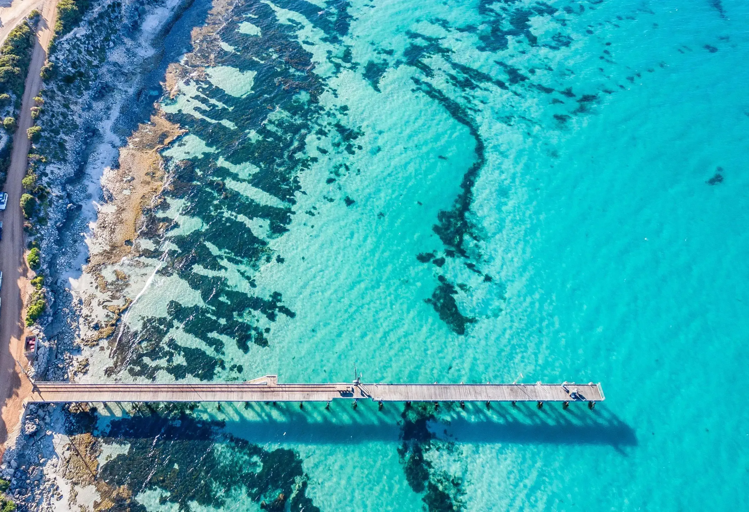 A jetty across a turquoise ocean along a beachside road with parked cars.