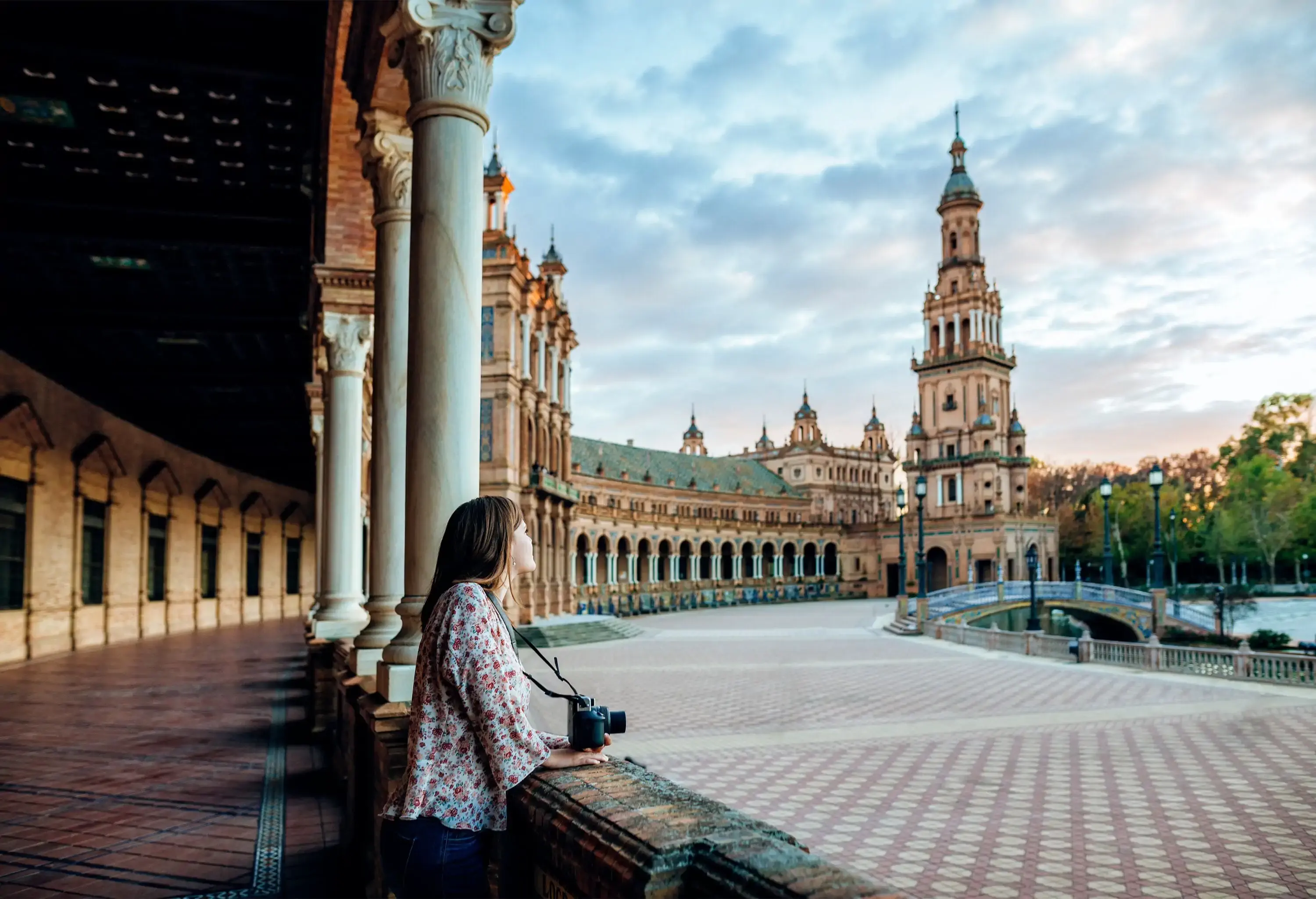 A woman with a camera leaning on the pillared hallways and gaze at the Renaissance tower of Plaza de España.