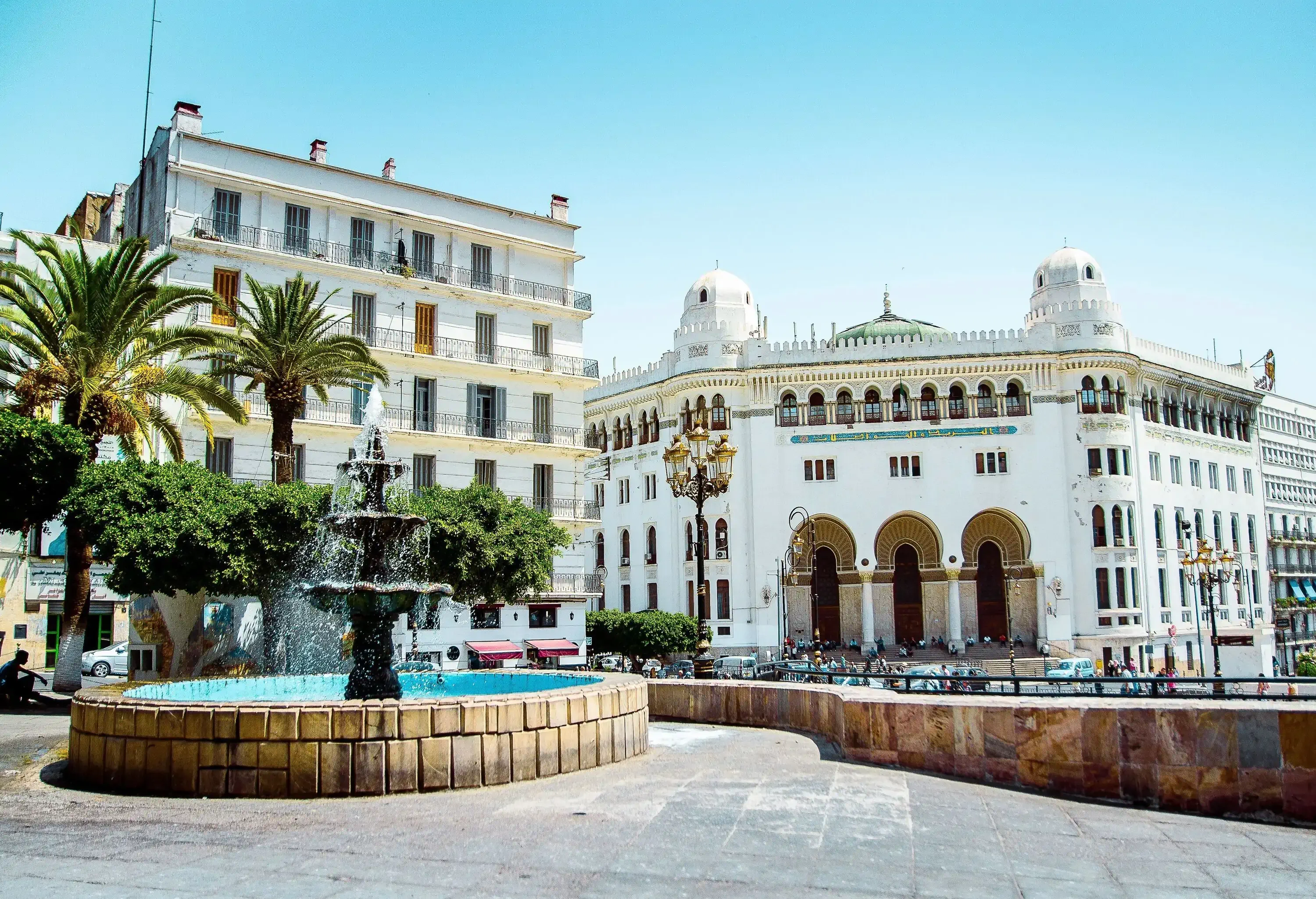 A Neo-Moorish post office building topped with domes line up with other structures alongside a bustling road near a fountain.
