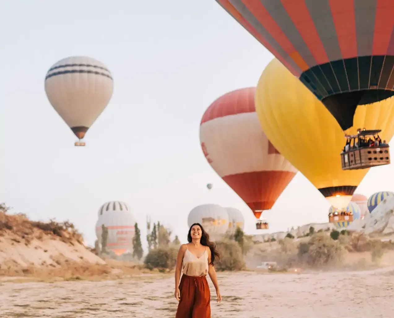Woman smiling while walking on the ground with colorful hot air balloons floating in the sky behind her.