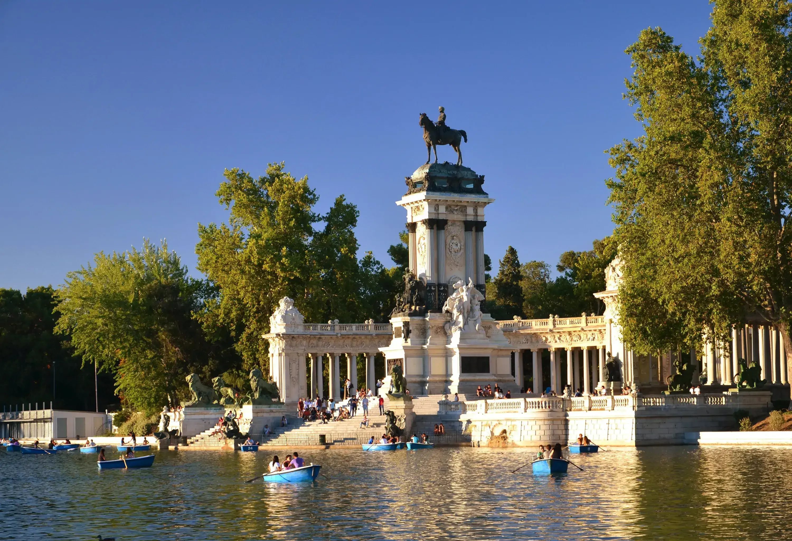 Sunlight illuminates a neoclassical monument and the surrounding trees in a public park, beside a bustling lake.