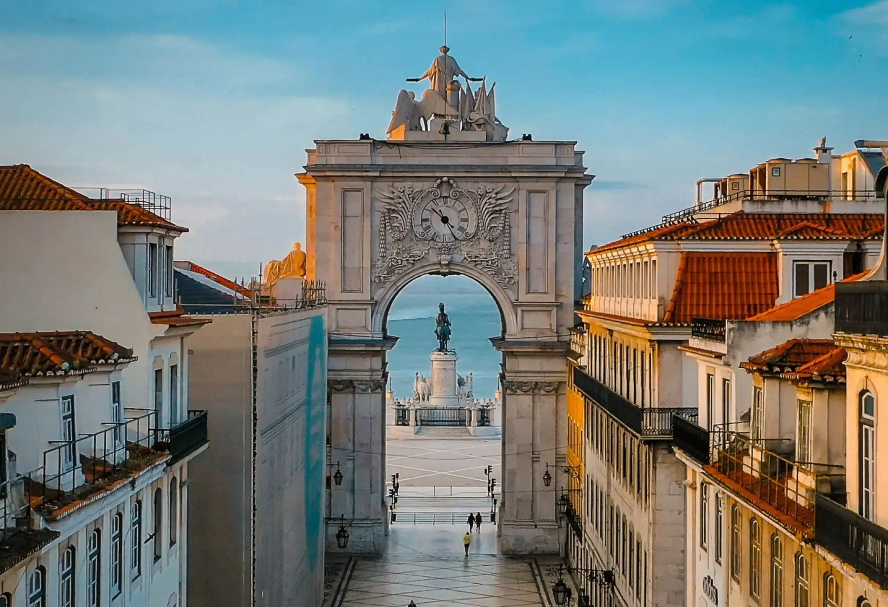 A long street leading to a grand triumphal arch, with classical buildings lining both sides and a large plaza visible beyond the arch.
