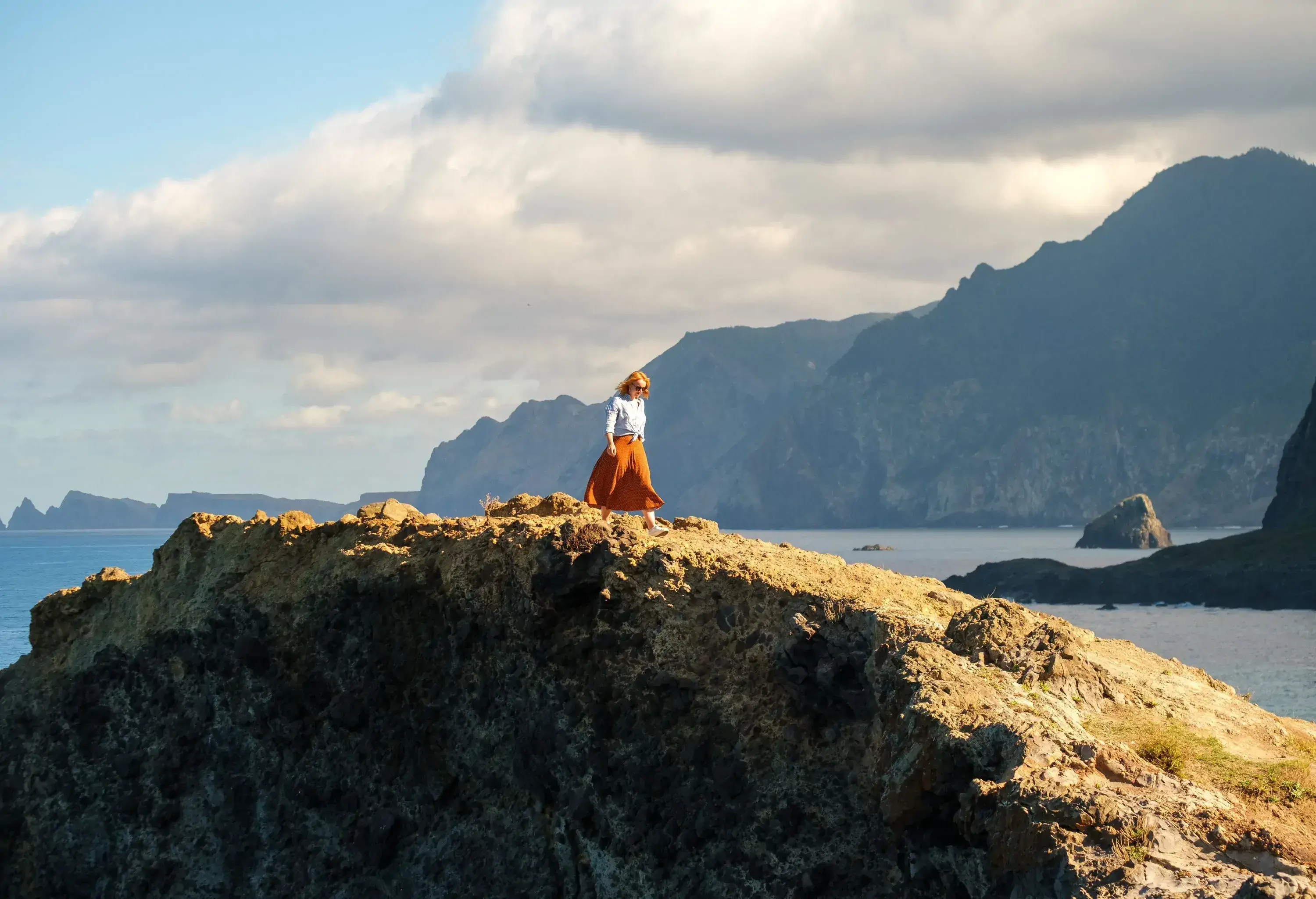 A woman walking down a seaside hill with large mountains in the backdrop.