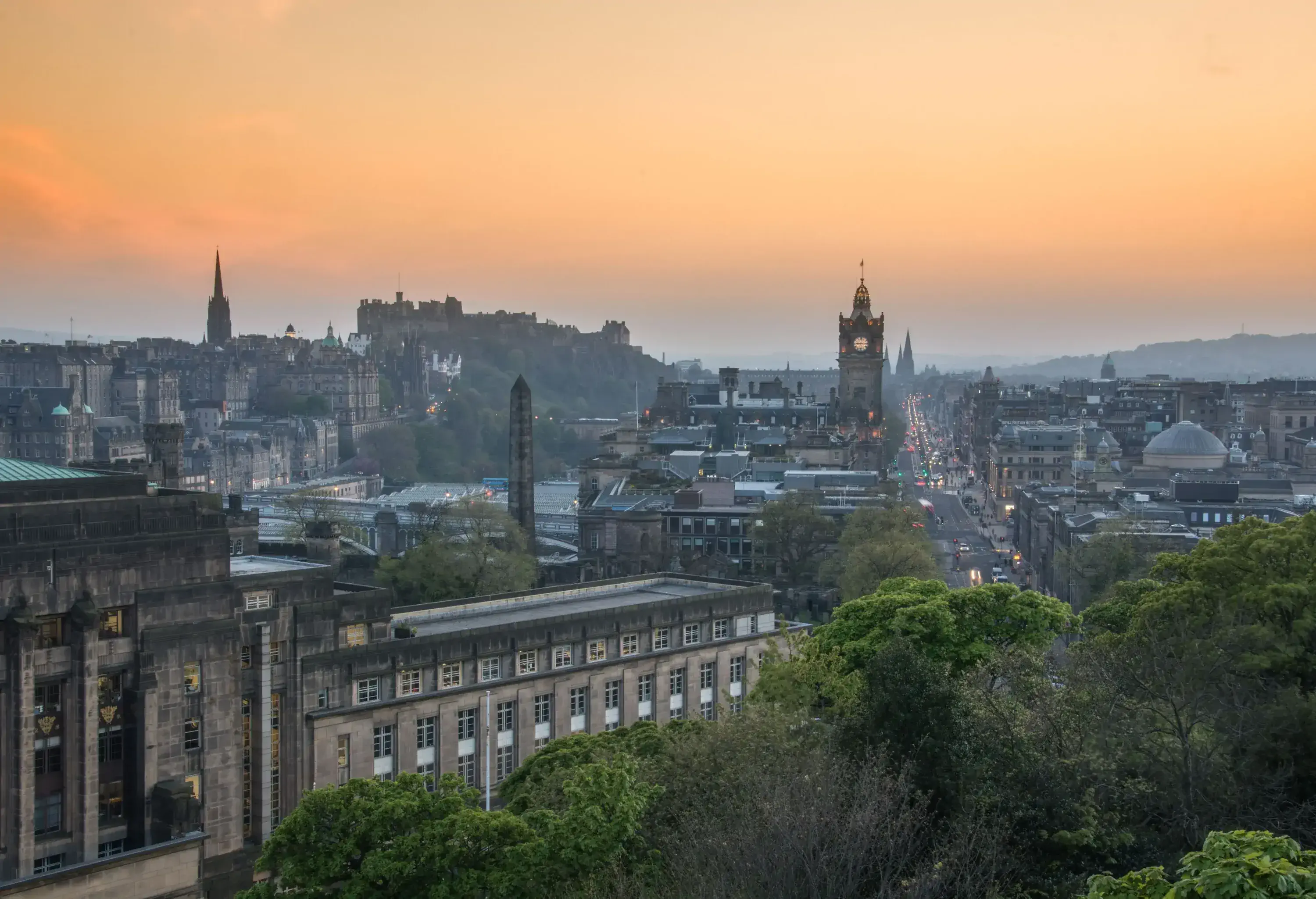 A clock tower rises above a cluster of old city buildings under an orange sky.