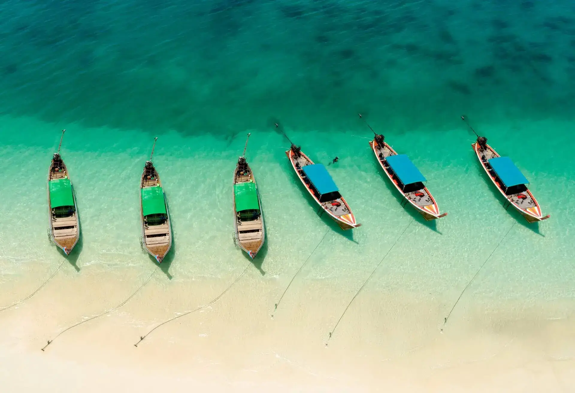 Six long-tail boats with red and blue roofs anchored on a pristine beach.