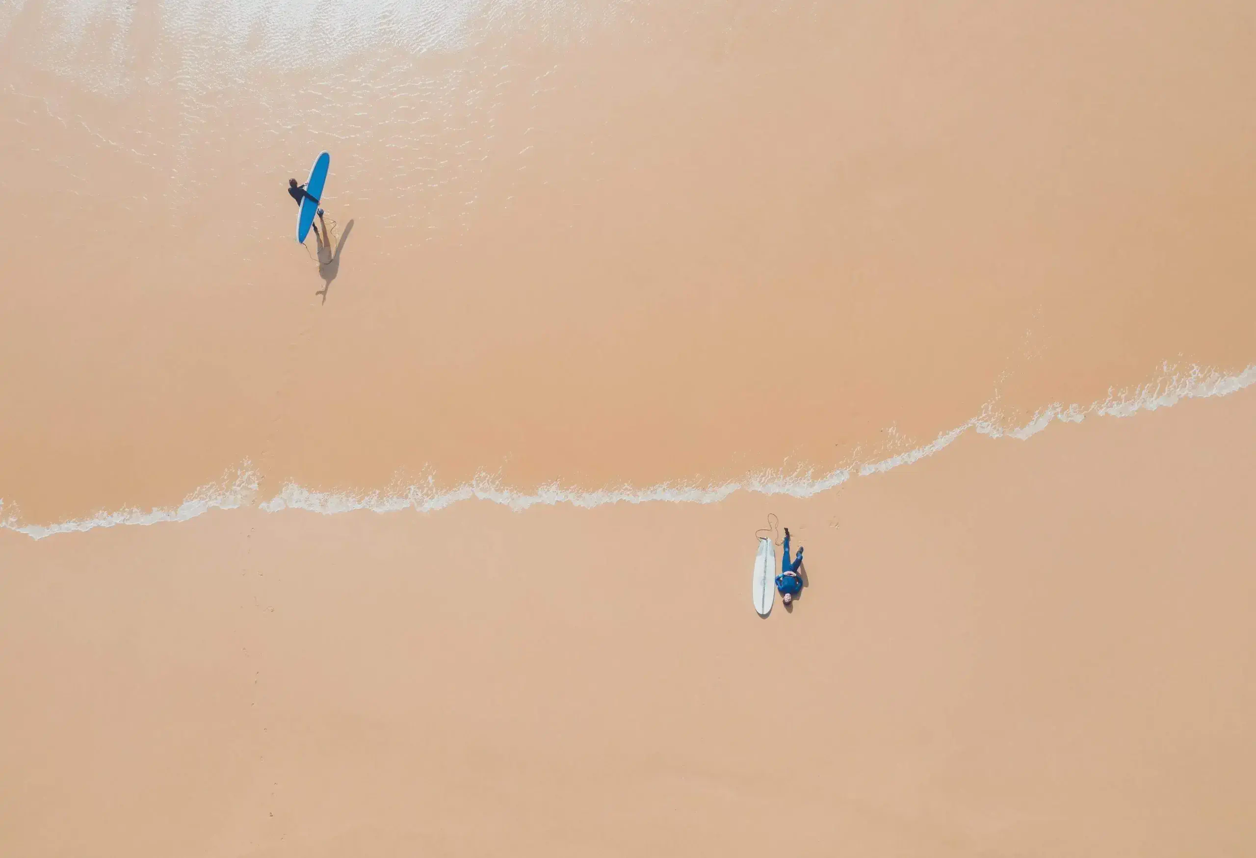 Surfers populate the beach, with one lounging casually beside their surfboard while another strides confidently across the sand, surfboard in tow, ready to conquer the waves.