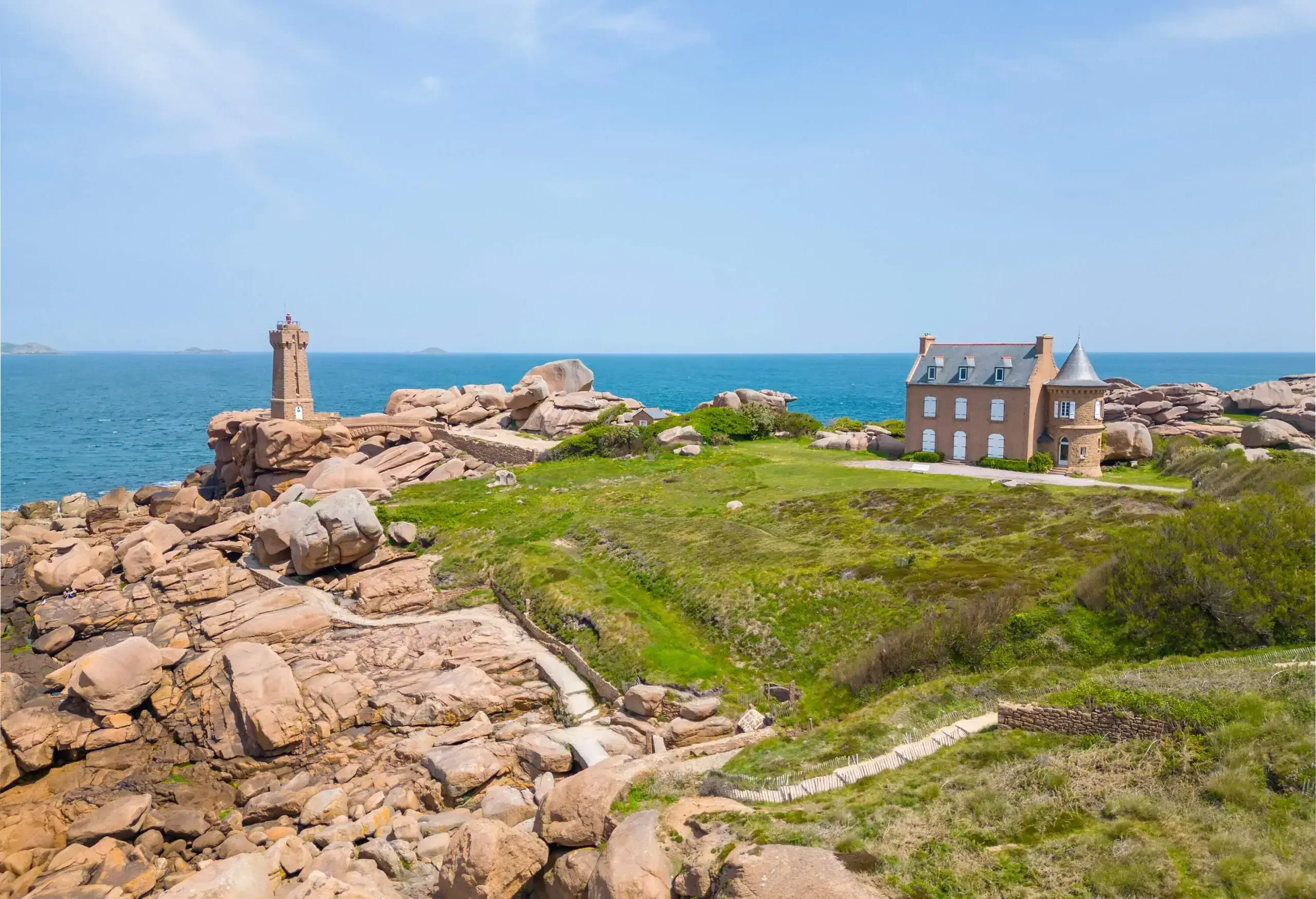 Wide angle view of a hill made or rocks and green grass, with a lighthouse by the sea and a large building