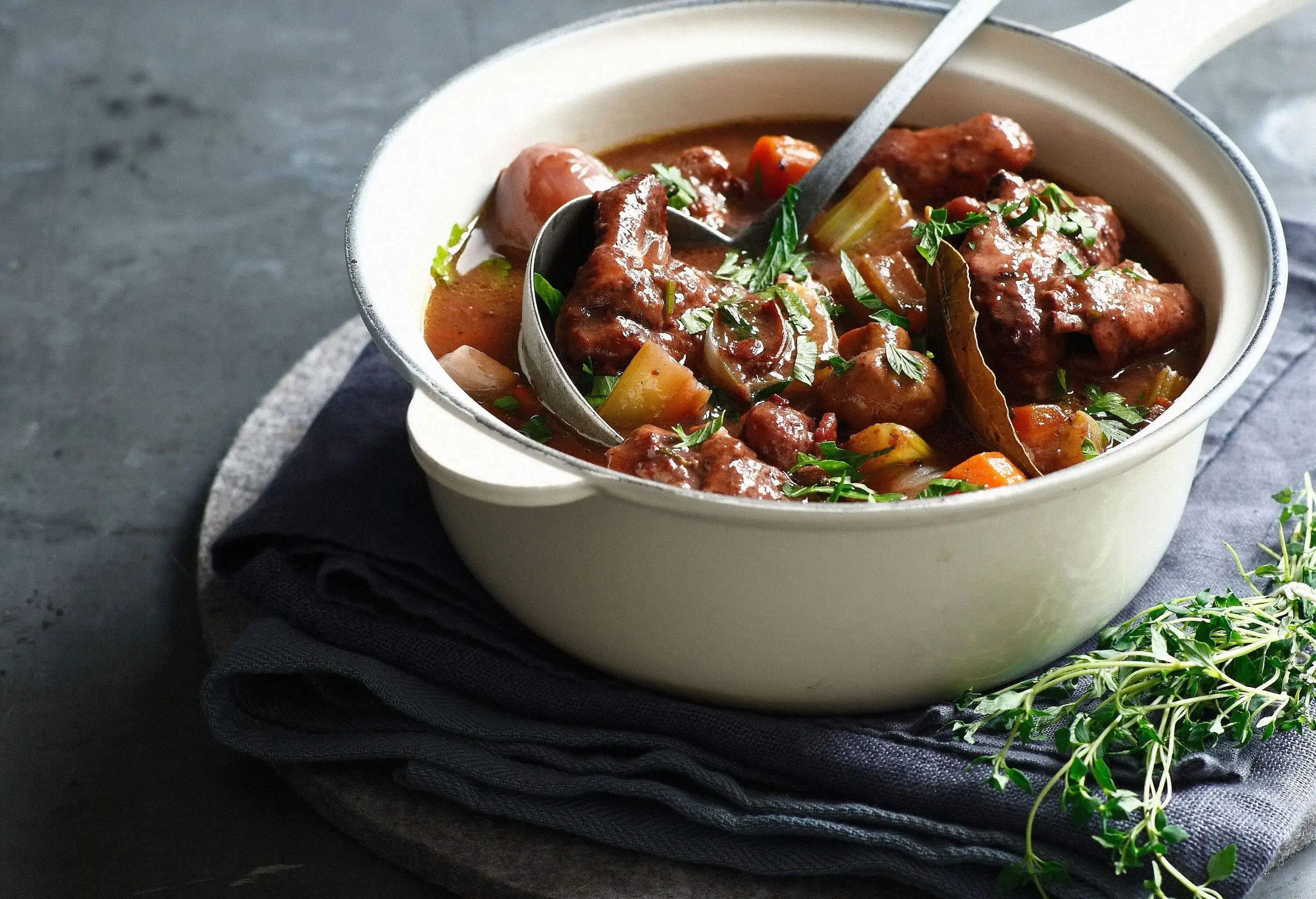 A bowl of savoury beef bourguignon topped on a wooden board with grey linen.