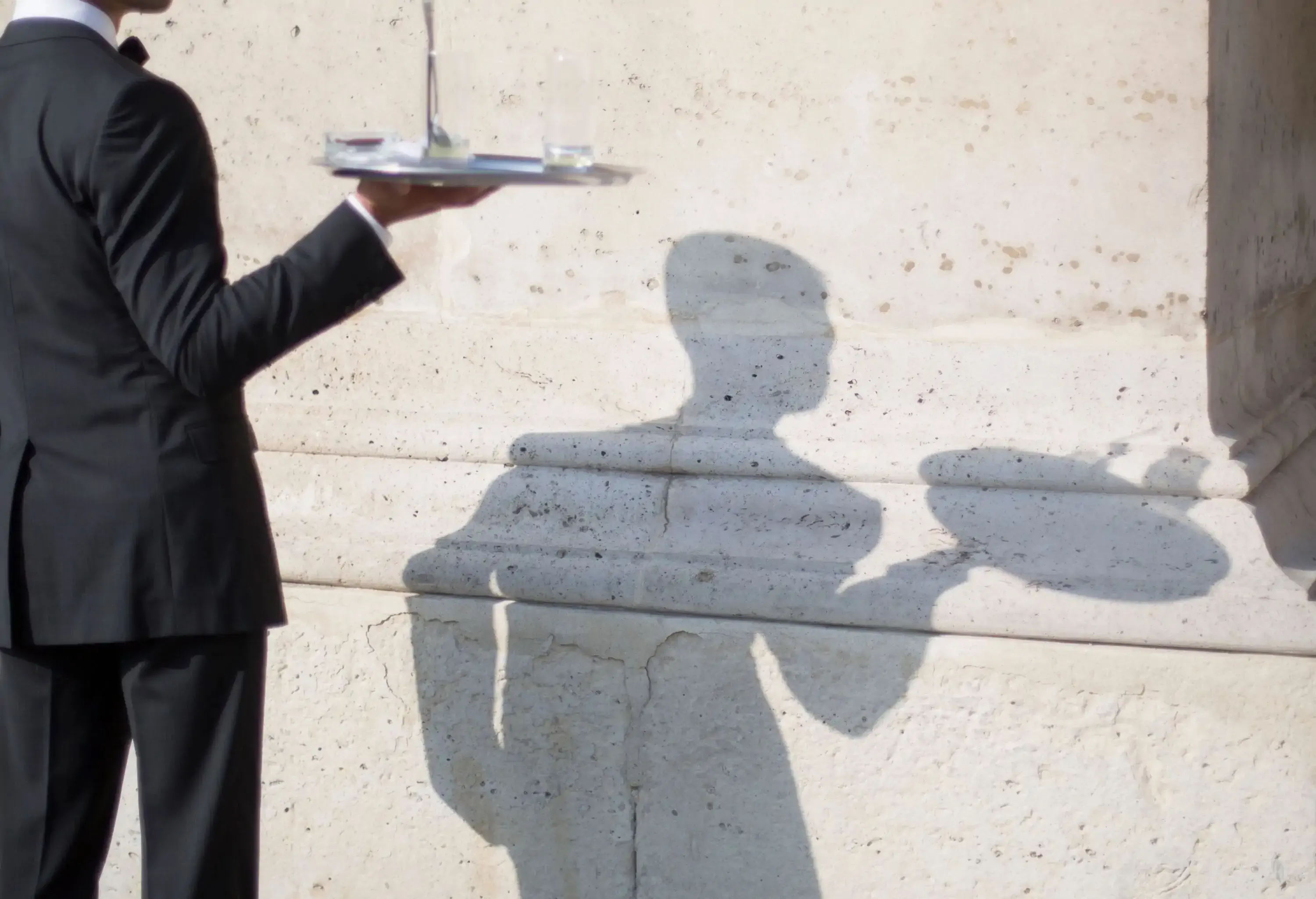 A waiter's shadow gracefully holds a tray, cast upon a textured concrete wall.