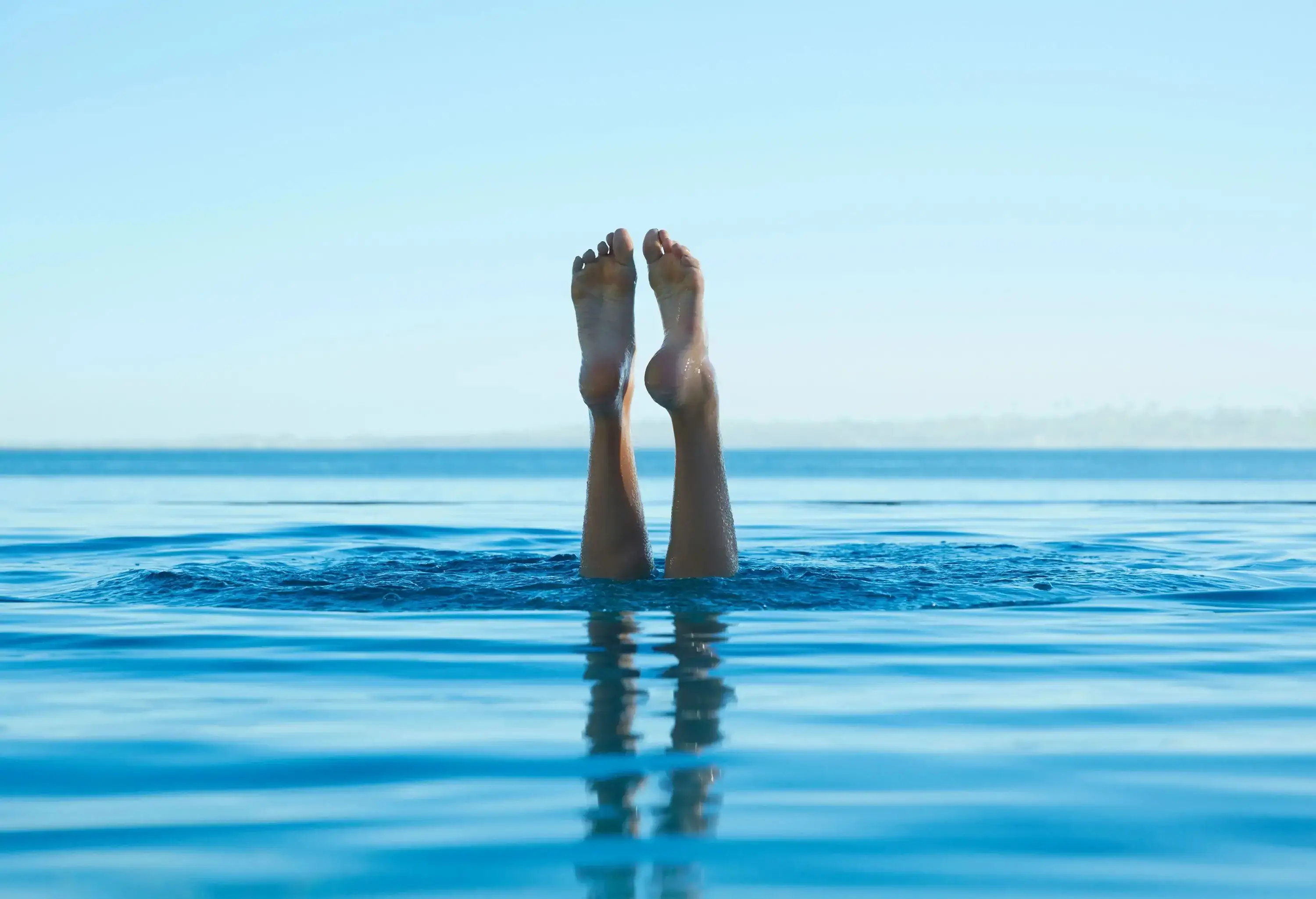 The feet protrude above the surface of dazzling turquoise waters, pointing to the cloudless sky.