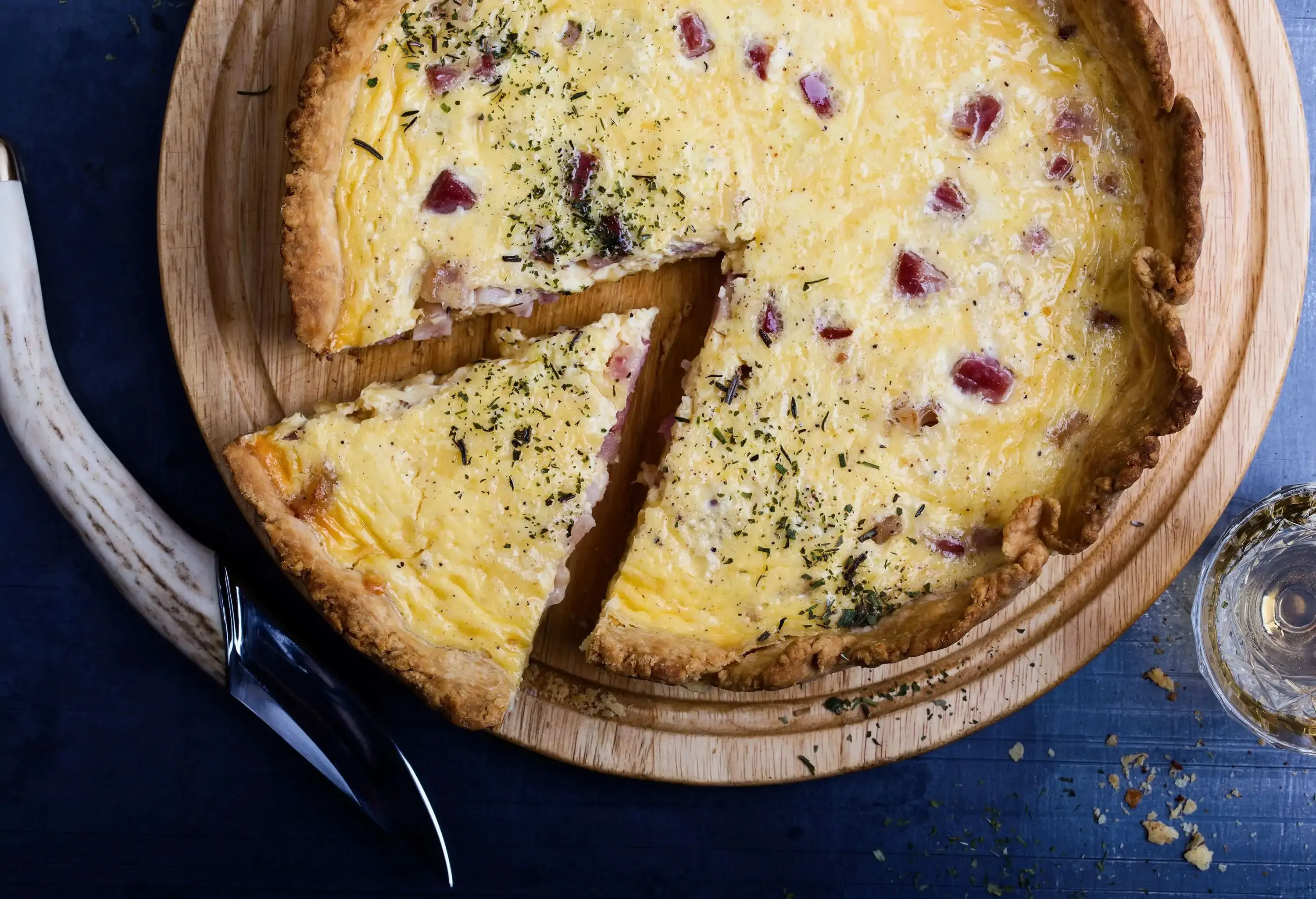 A freshly baked quiche stuffed with bacon and cheese rests on a wooden cutting board, along with a chunk of the quiche that has been sliced.