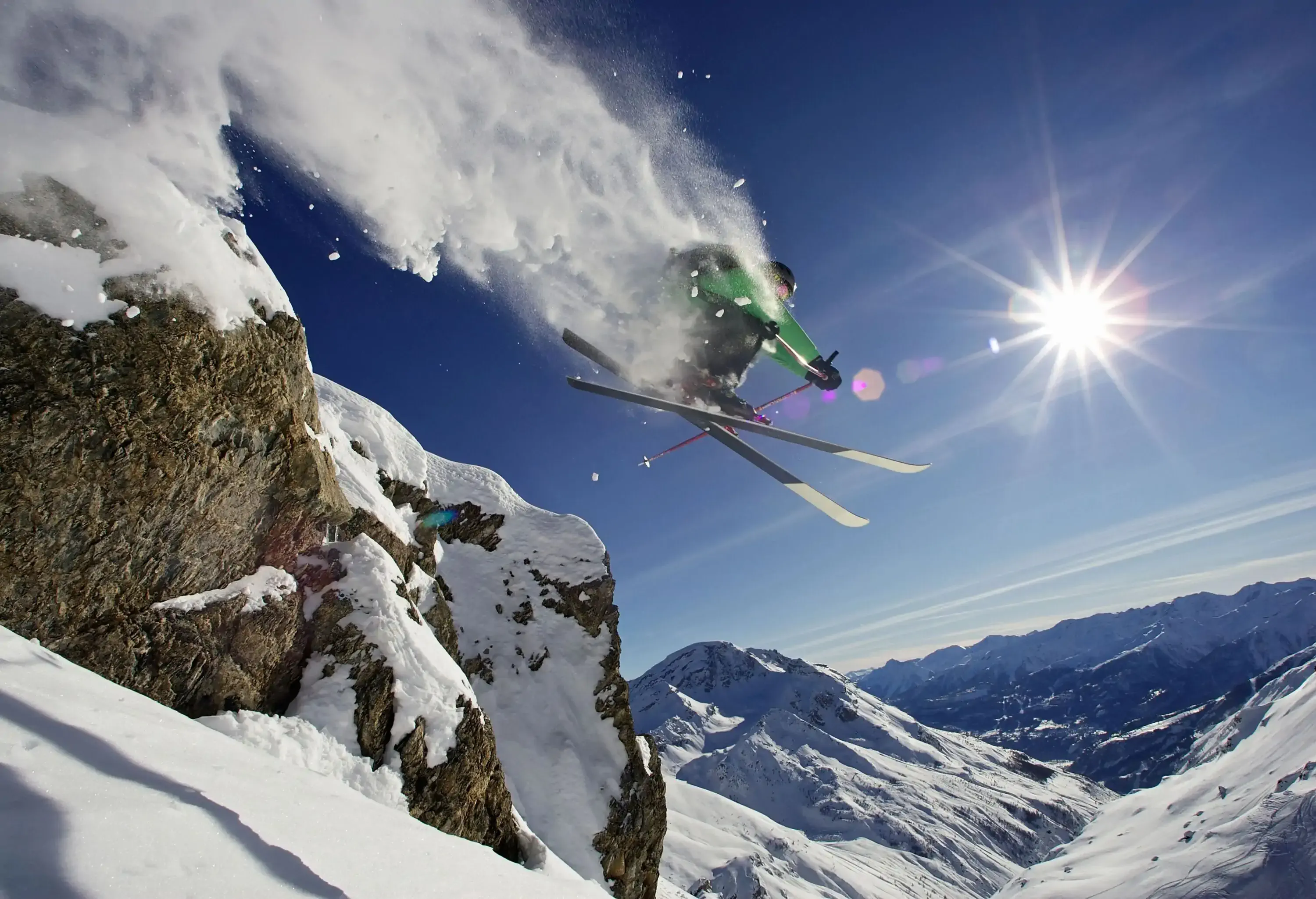 A skier flying in the air, spraying snow as he goes, over a backdrop of snowy mountains.