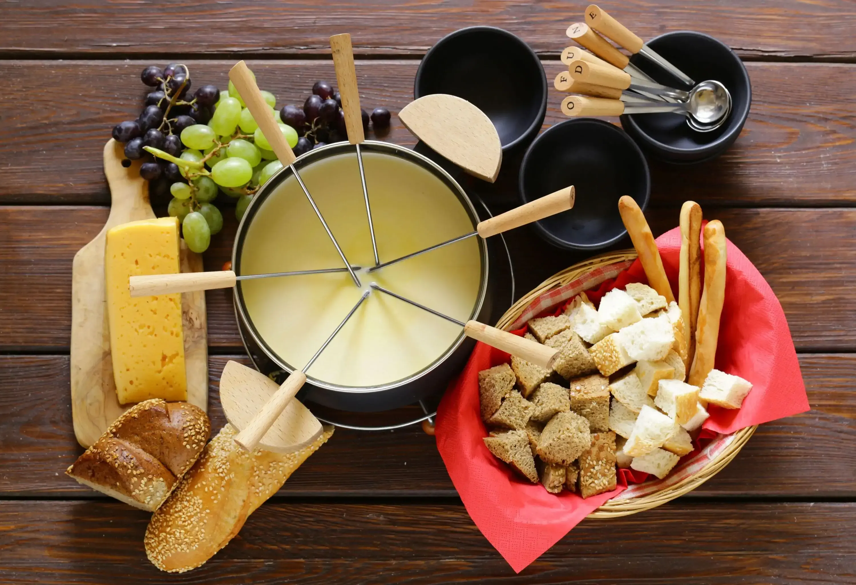 Melted cheese and wine on a pot with long-stemmed forks served with grapes, loaves, and a basket of sliced bread.