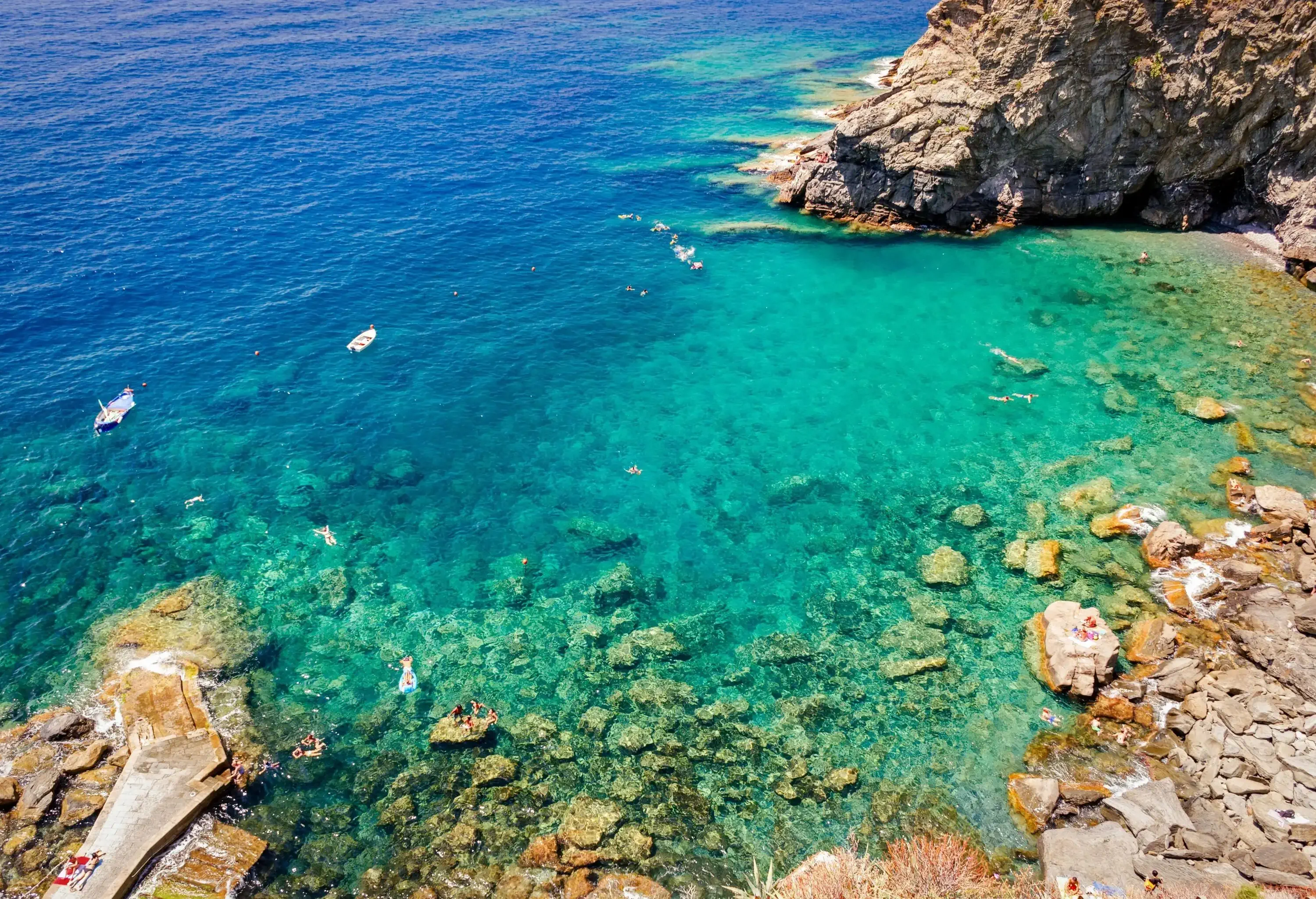 A rocky beach with some tourists swimming in its crystalline turquoise water and two white moored boats along a rugged cliff.