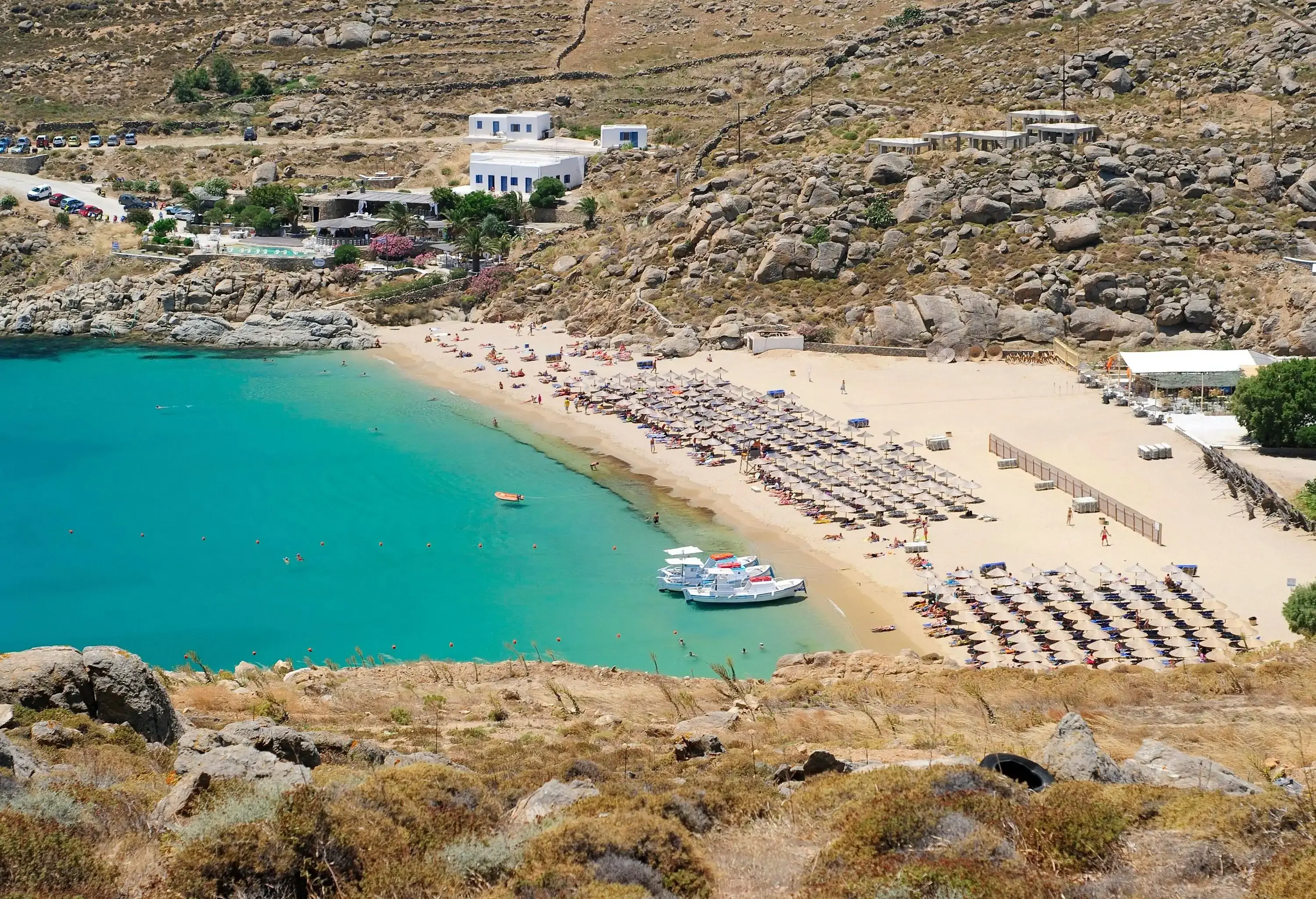 A sun-kissed beach with rows of colourful parasols and sun loungers, filled with people enjoying the warm weather, all set against the stunning backdrop of rocky hills with a white building complex perched atop.