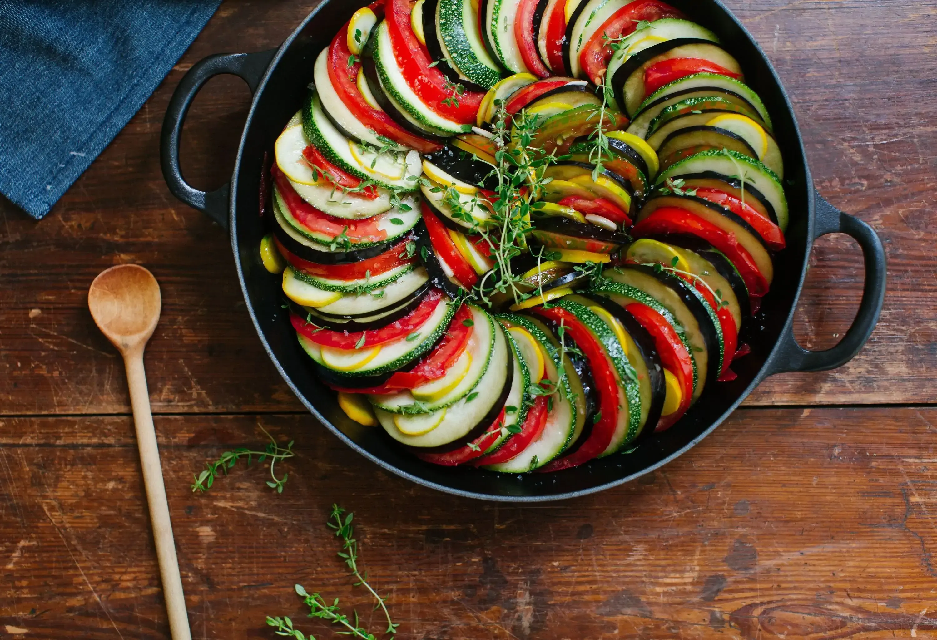 A rustic pot brimming with a colourful medley of vegetables, including eggplants, zucchini, and tomatoes, cooked to perfection in a flavorful ratatouille, accompanied by a wooden ladle, set against a charming wooden background.