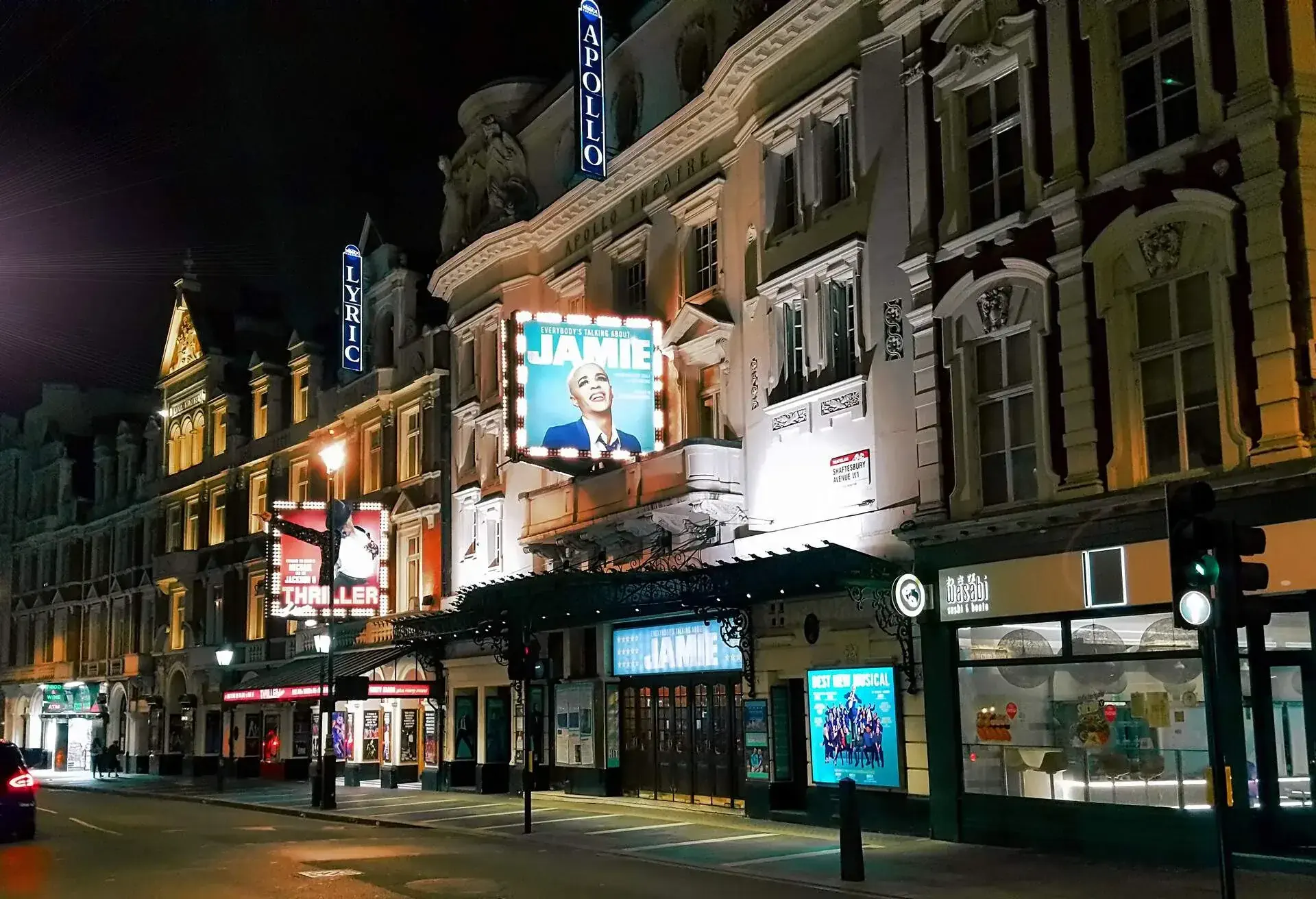 A major road's sidewalk illuminated by theatres posters and business signs.