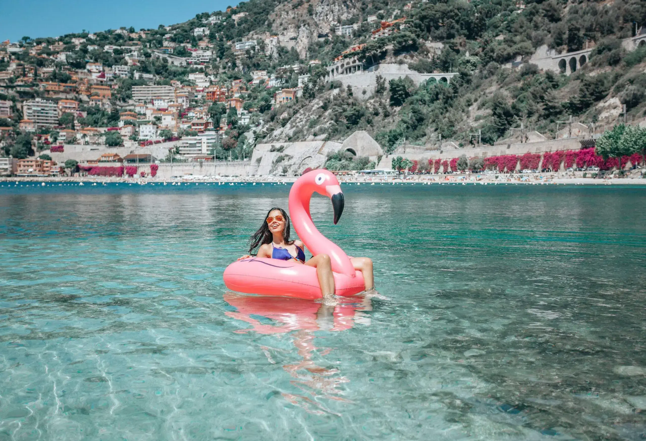 A woman in blue swimwear sits on a flamingo floater over the sea against the backdrop of a colourful coastal city on a hilly island.