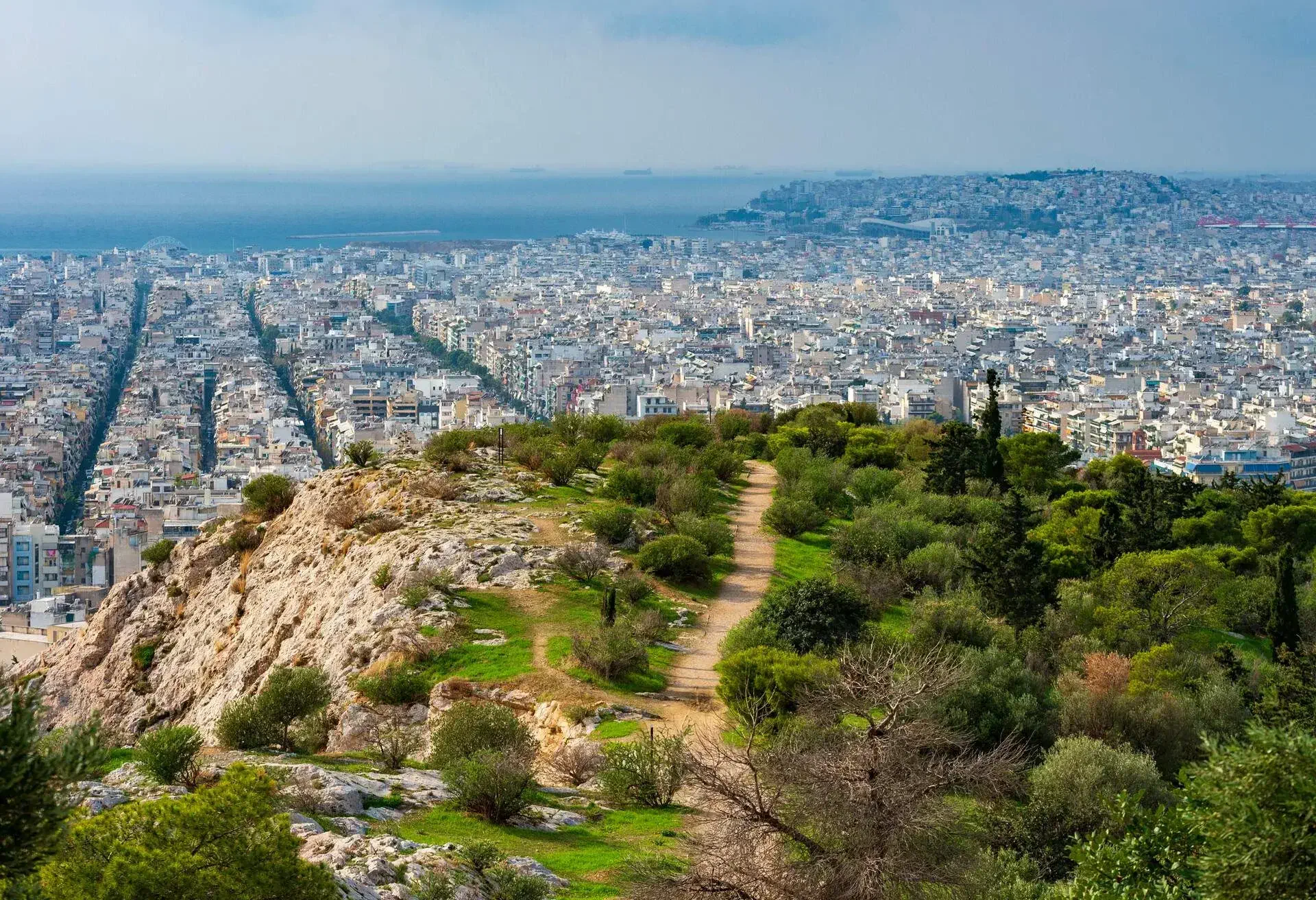 View of the Greek capital city of Athens from Philopappou Hill. A path winds between trees and stones down into the city. View of straight streets from a height.