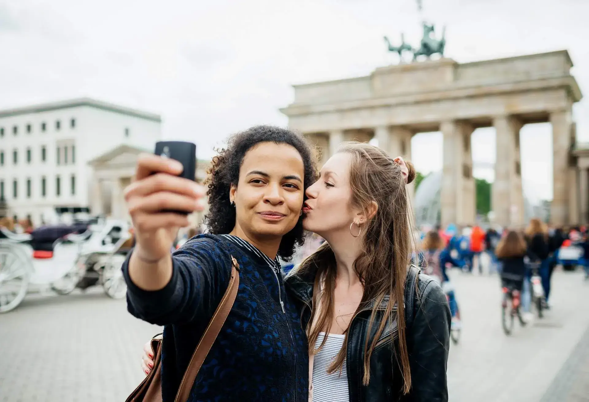 A young couple stops for a moment to take a selfie at a local historical site, one tenderly planting a kiss on the other's cheek.