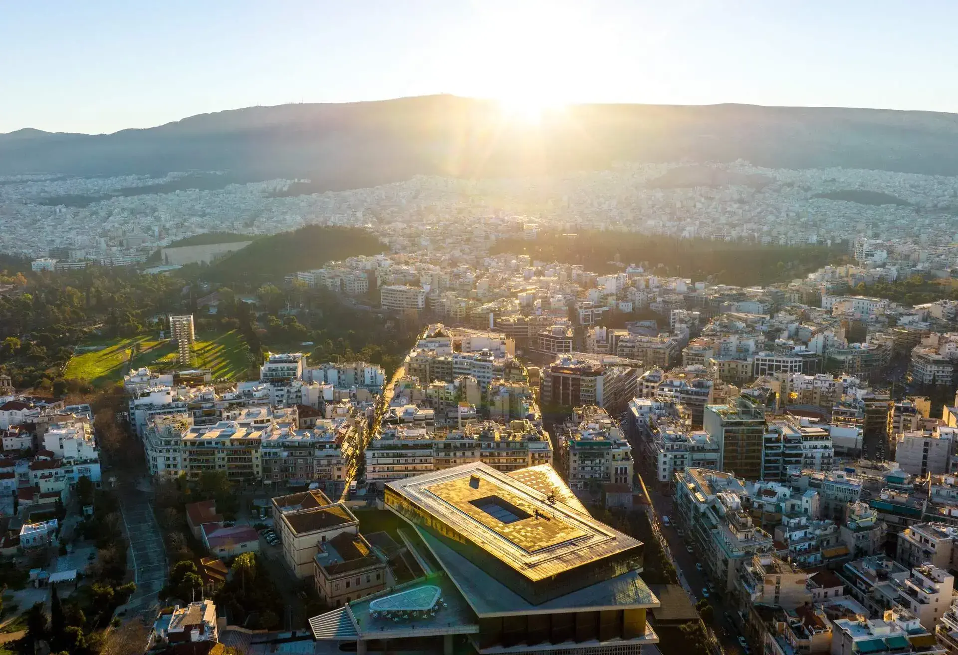 This is an aerial photo of the sunrise over Hymettus mountain in Athens, Greece. The large building immediately below is the New Acropolis museum.