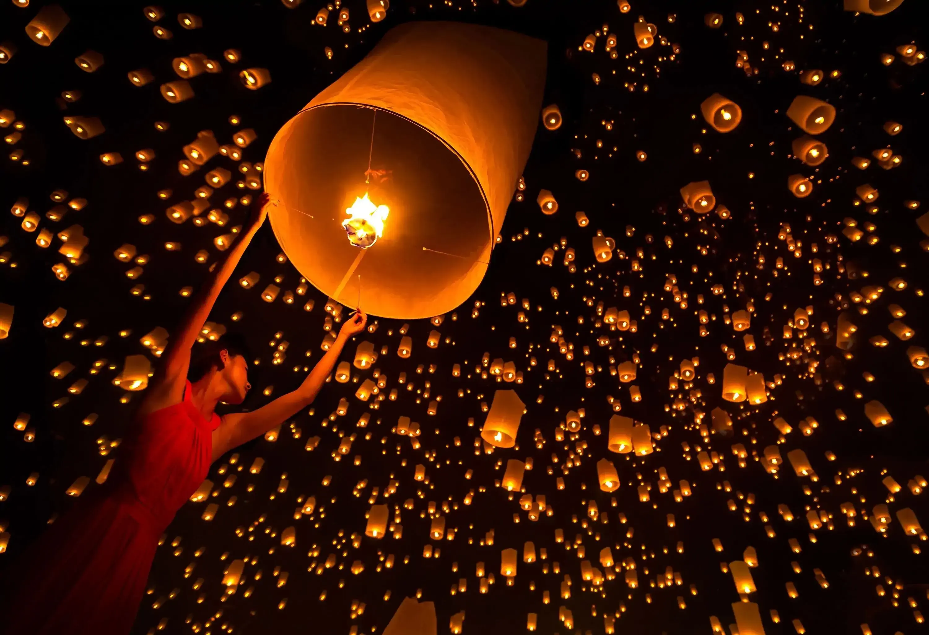 A woman releases a cylindrical sky lantern, with views of a cluster of sky lanterns floating in the sky.