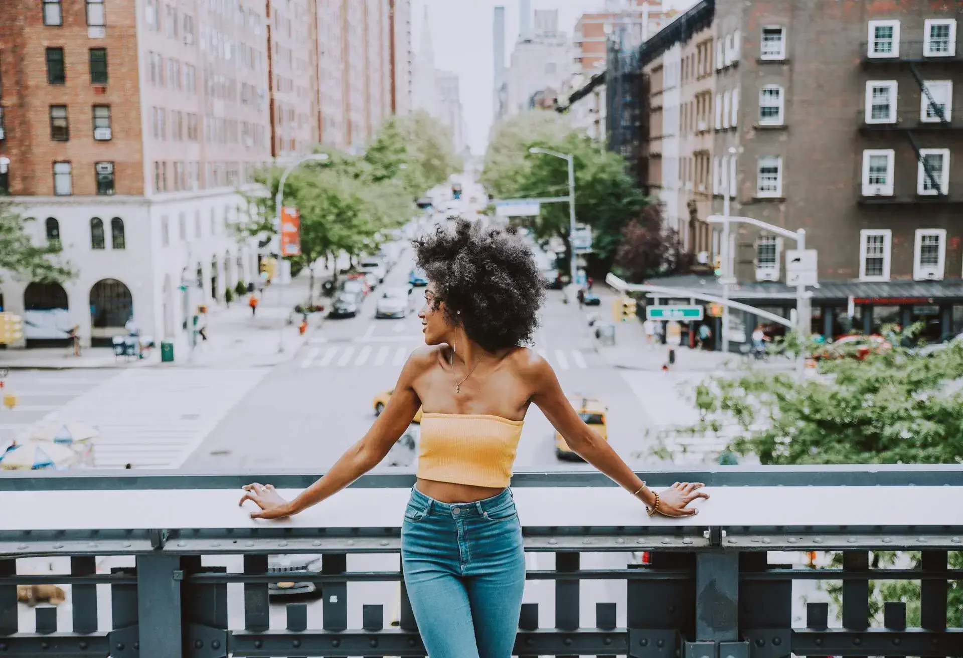 A curly-haired woman in a yellow tube top and denim jeans leans on a bridge railing as she looks sideways.