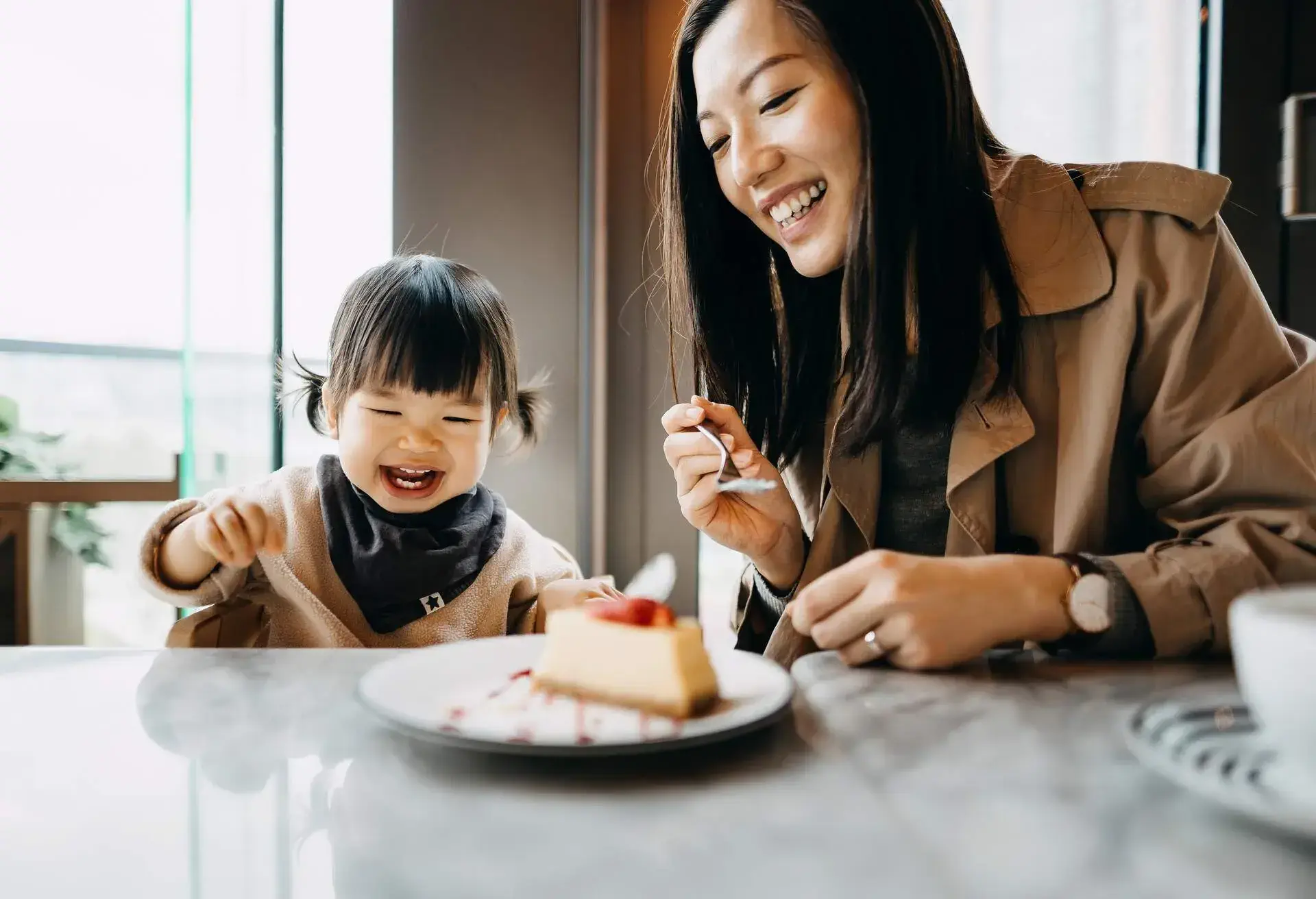 Mother and little daughter sharing a piece of cake in the cafe, both of them are smiling joyfully