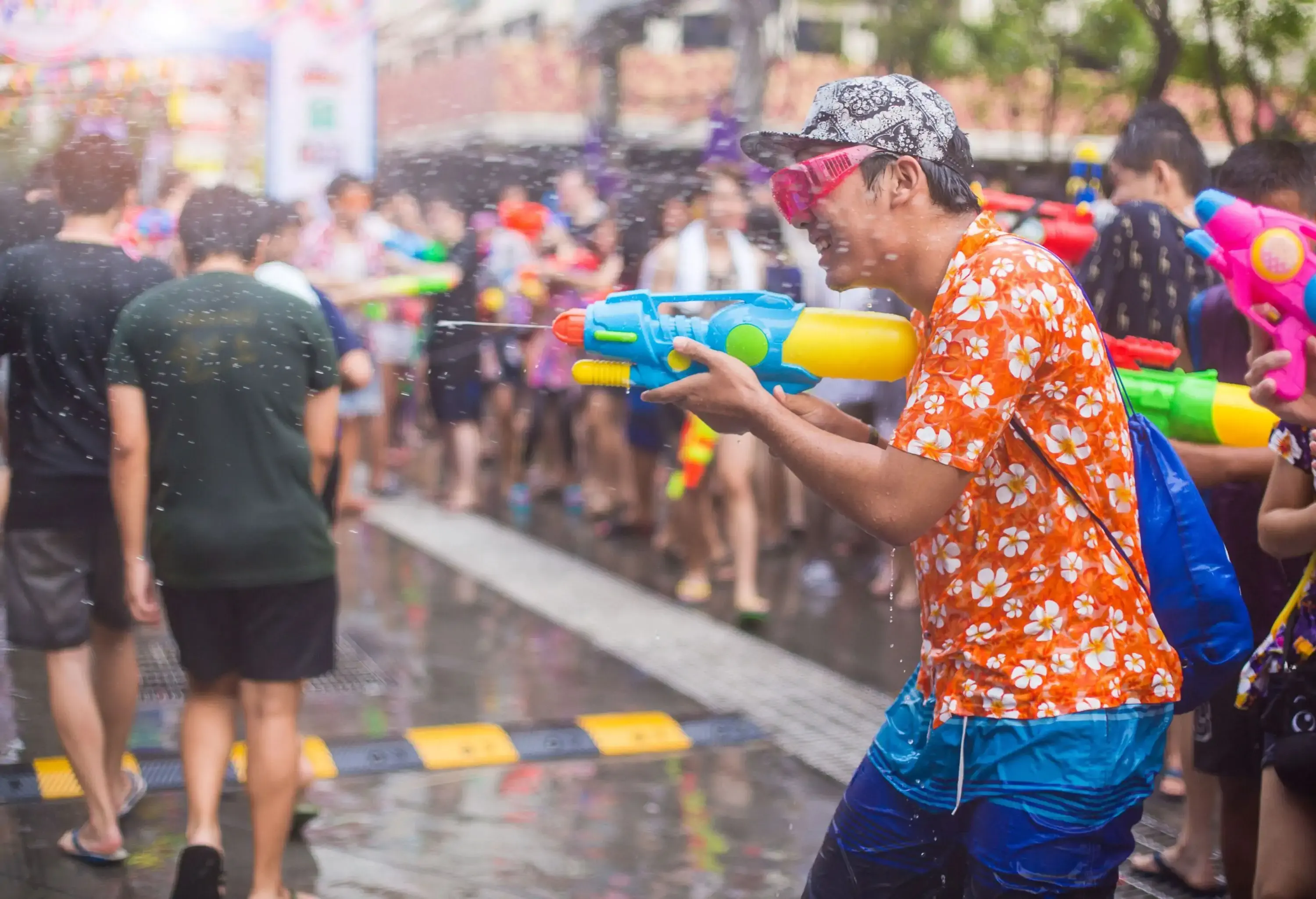 Man wearing an orange floral shirt uses a colourful water gun to blast water at people.