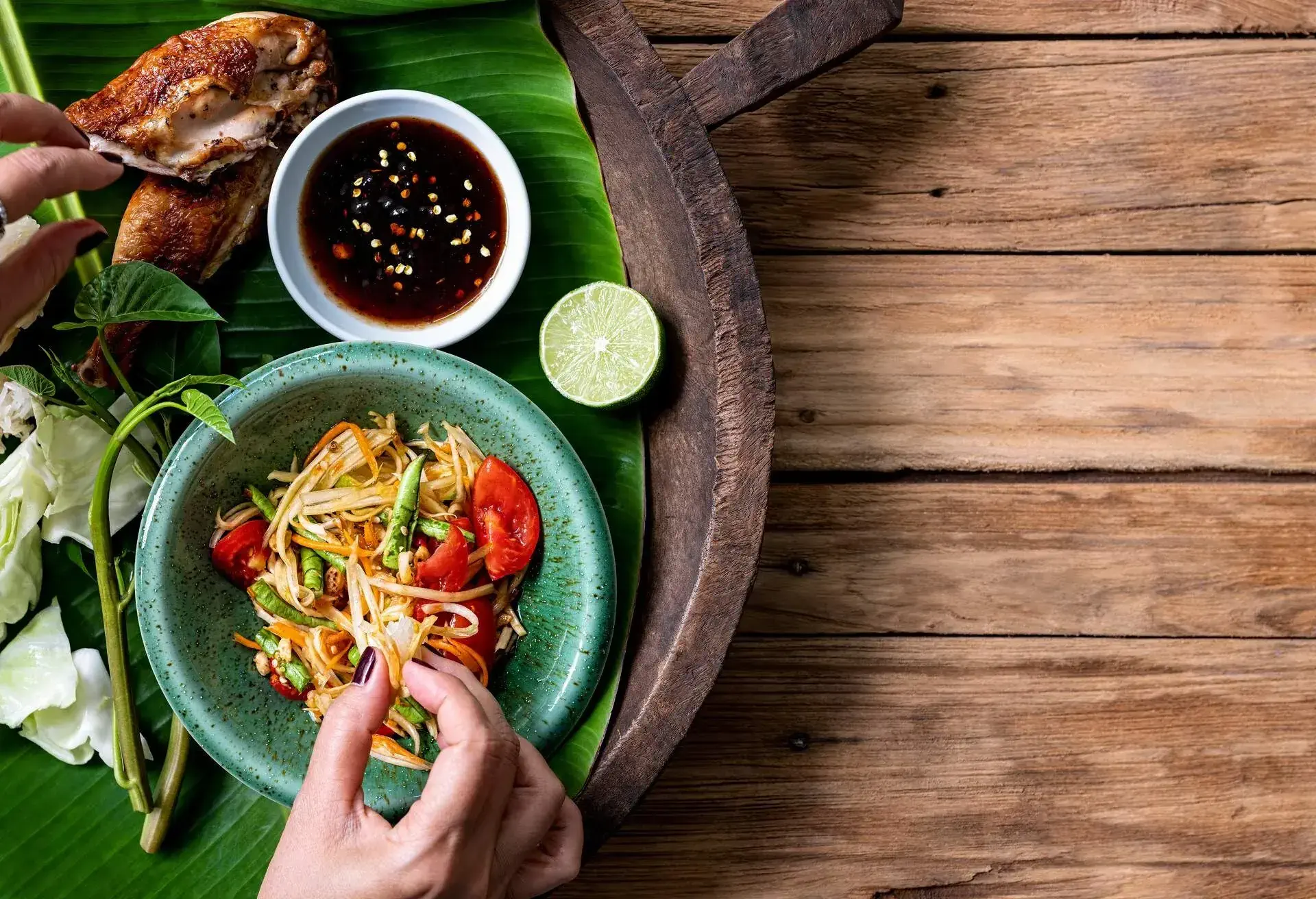 Thai lady eating traditionally with her hands, fresh world famous Som Tam (papaya salad) with BBQ chicken, sticky rice and raw salad vegetables on an old wooden table background. Good copy space to the right of the image.