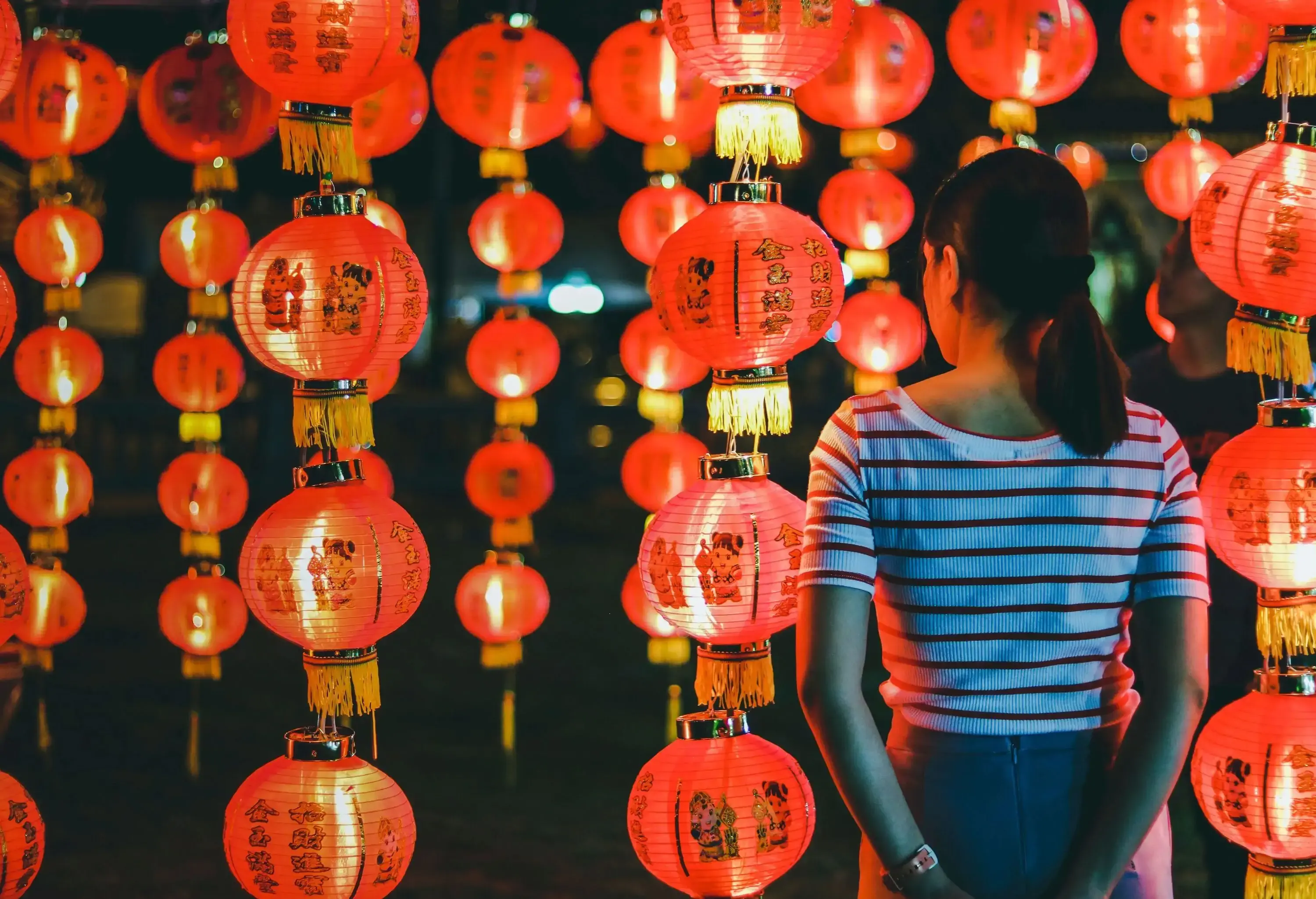 A woman in a striped shirt stands amongst a series of bright red lanterns.