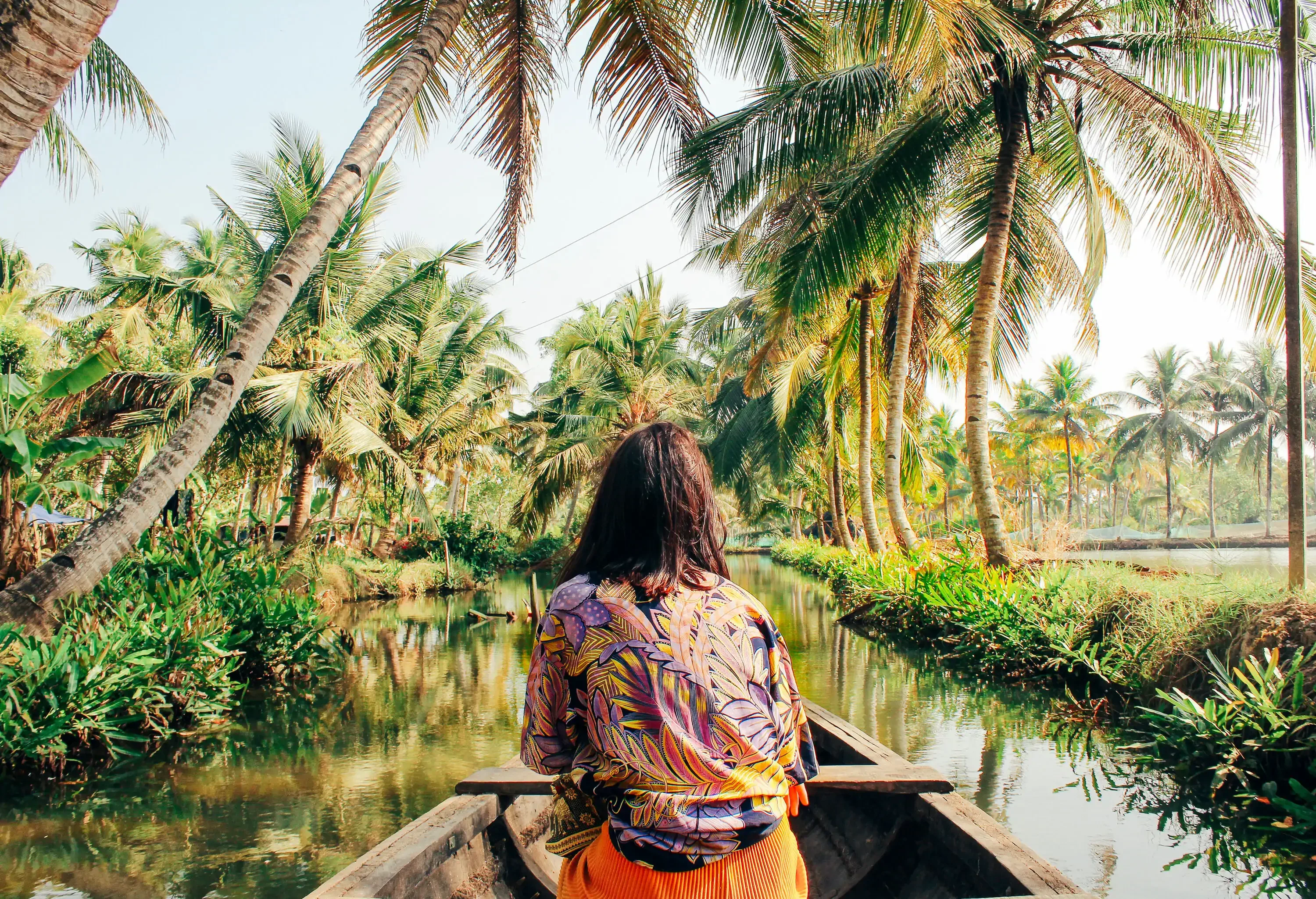 A woman in a colourful printed dress sits on a wooden boat that cruises across a backwater under the canopy of palm trees.