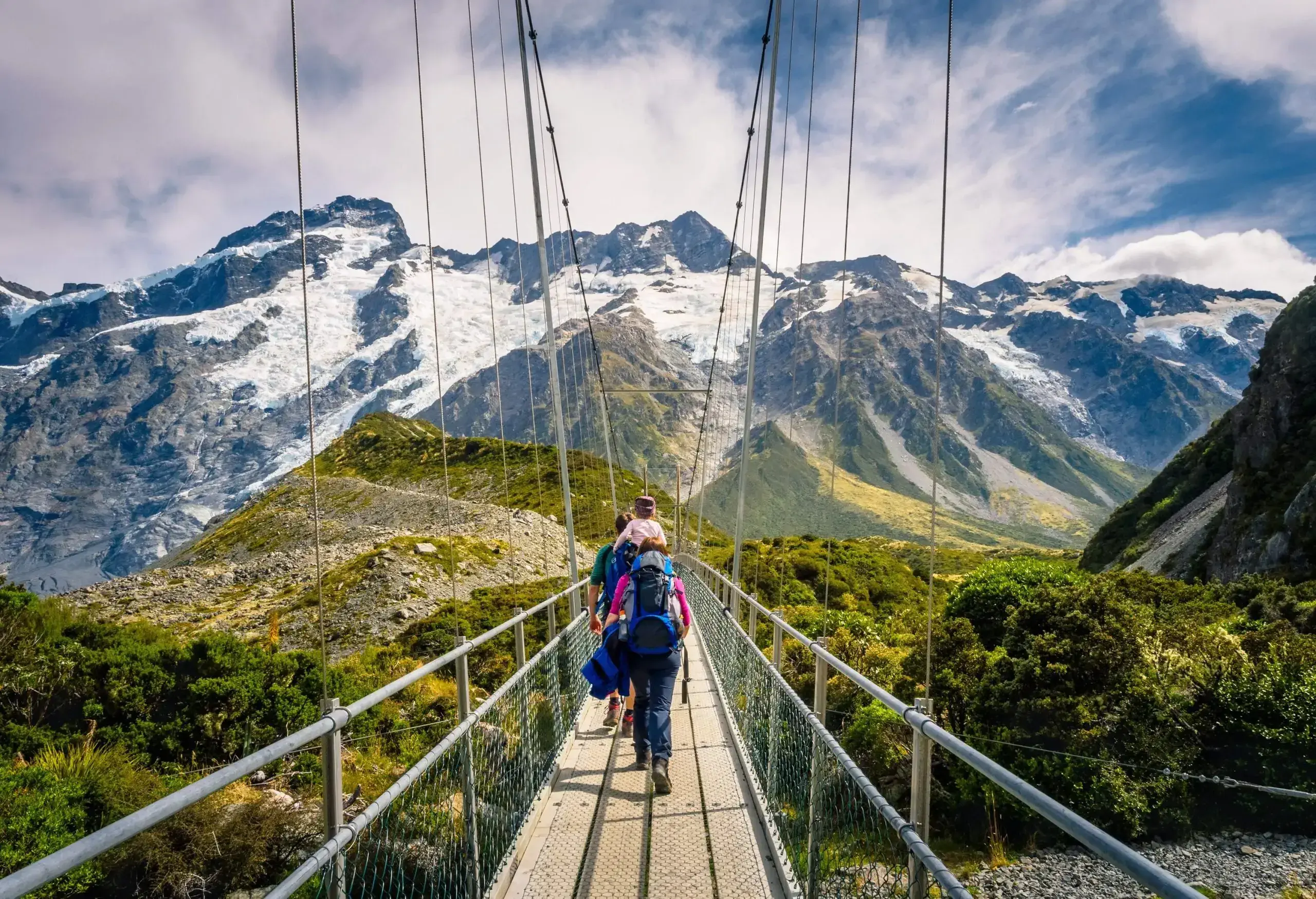 A family crosses a suspension footbridge heading toward the spectacular steep snowy mountains.