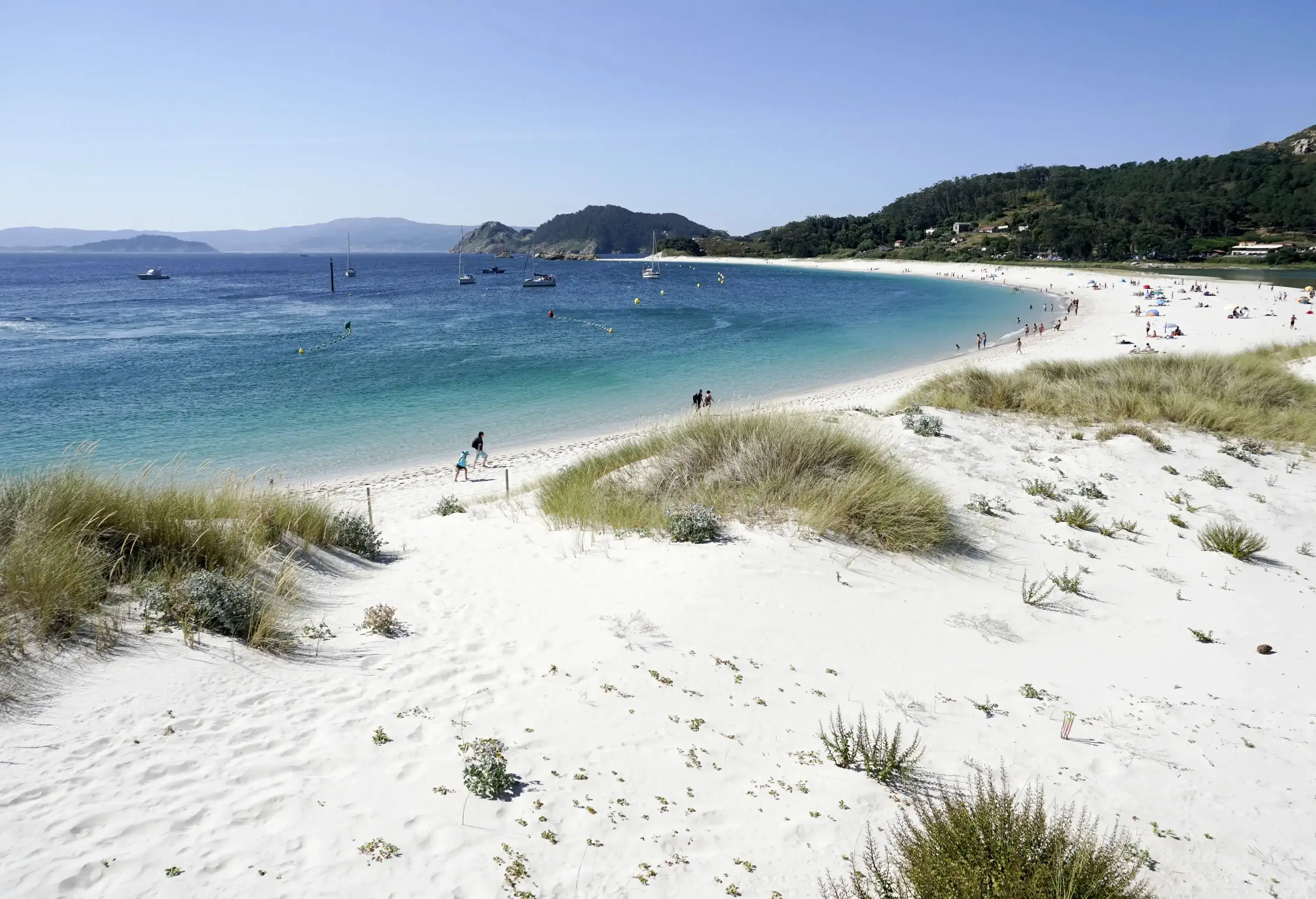 A white sand beach with boats and people is seen at the base of a lush, forested hill.