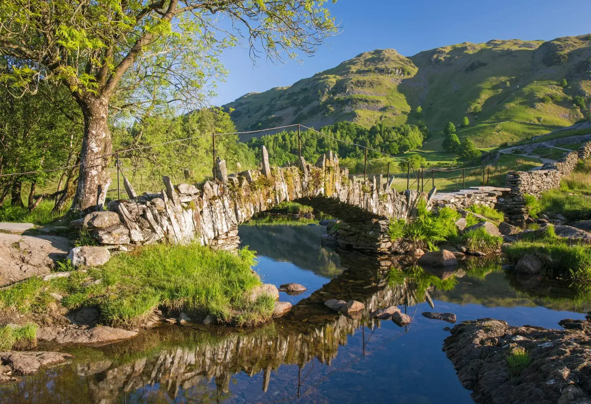 Slater's Bridge reflected in the tranquil waters of the River Brathay, early morning, Little Langdale, near Ambleside, Lake District National Park, Cumbria, England, UK, Europe. Famed for the beauty of its lakes and mountains, the Lake District is a popular holiday destination in northwest England. Lying entirely within the county of Cumbria, it became the UK's second National Park in 1951 and was designated a UNESCO World Heritage Site in 2017. Slater's Bridge is a former packhorse bridge which dates back to the 16th century.
