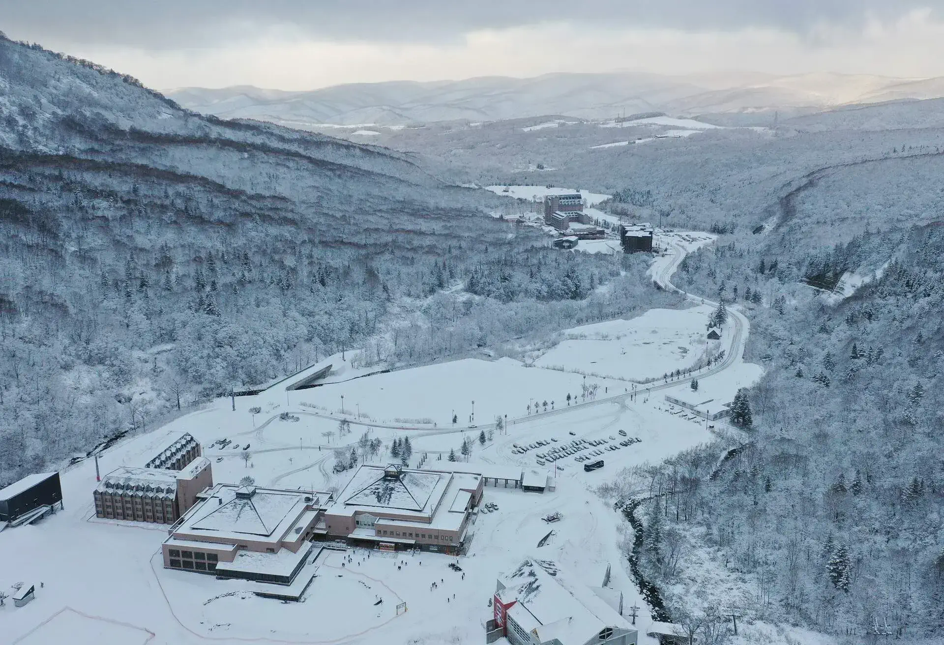 Snow-covered buildings of a ski resort bordered by a frozen forest on the hills.