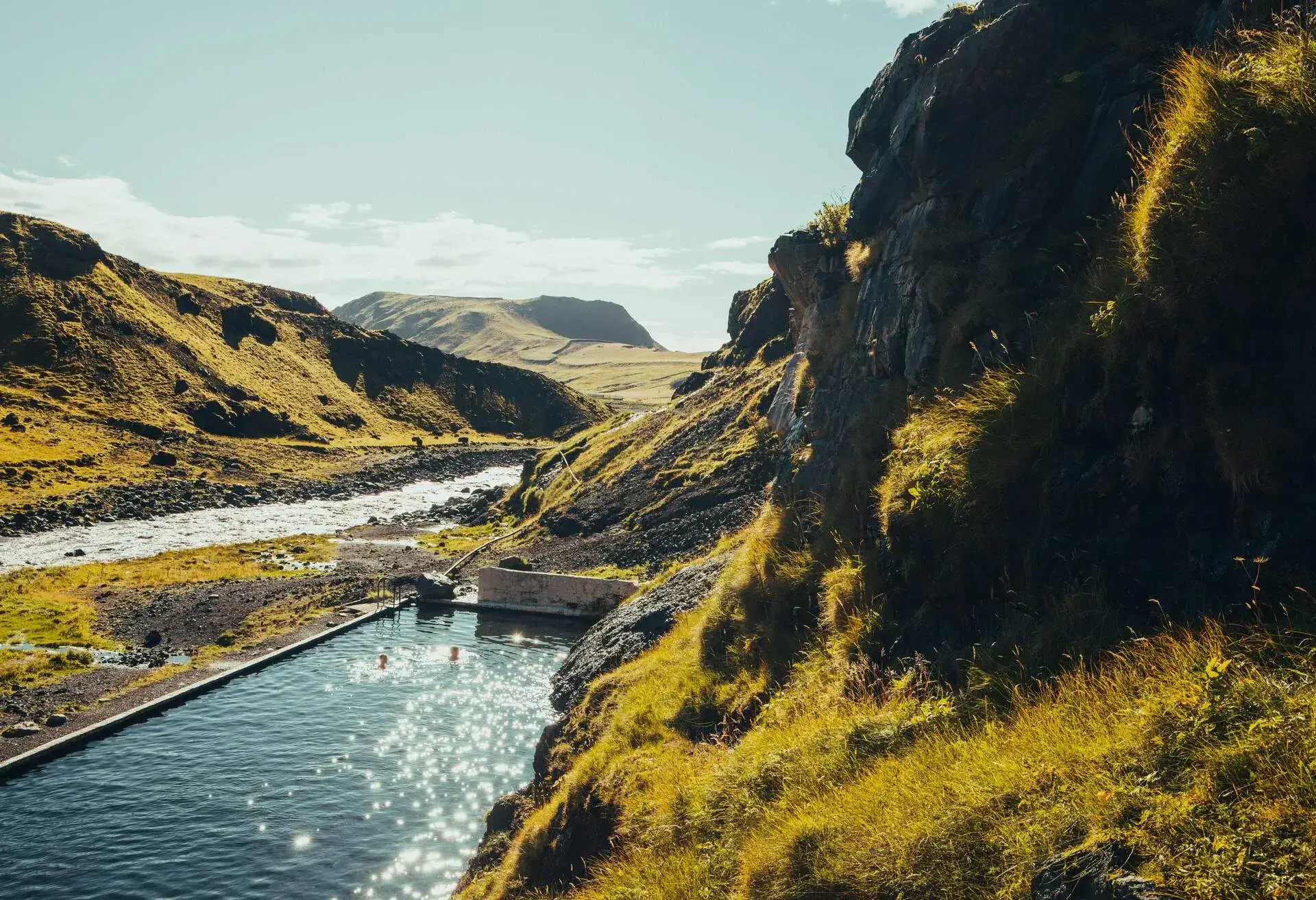 Female couple swimming in the Seljavallalaug Swimming Pool with bright sun reflecting light in the water surface, and the landscape with green vegetation covering the volcanic landscape in Iceland.