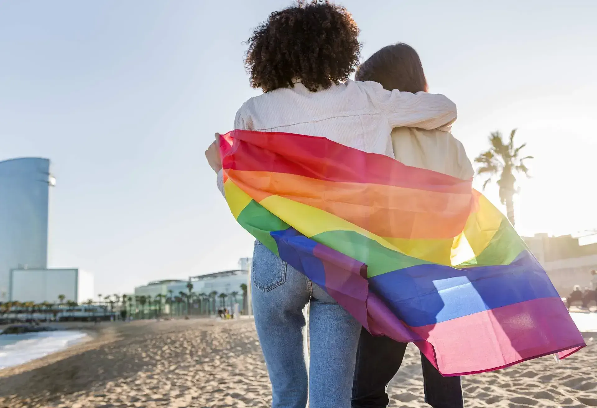 Two people shrouded in a Pride flag on the beach.