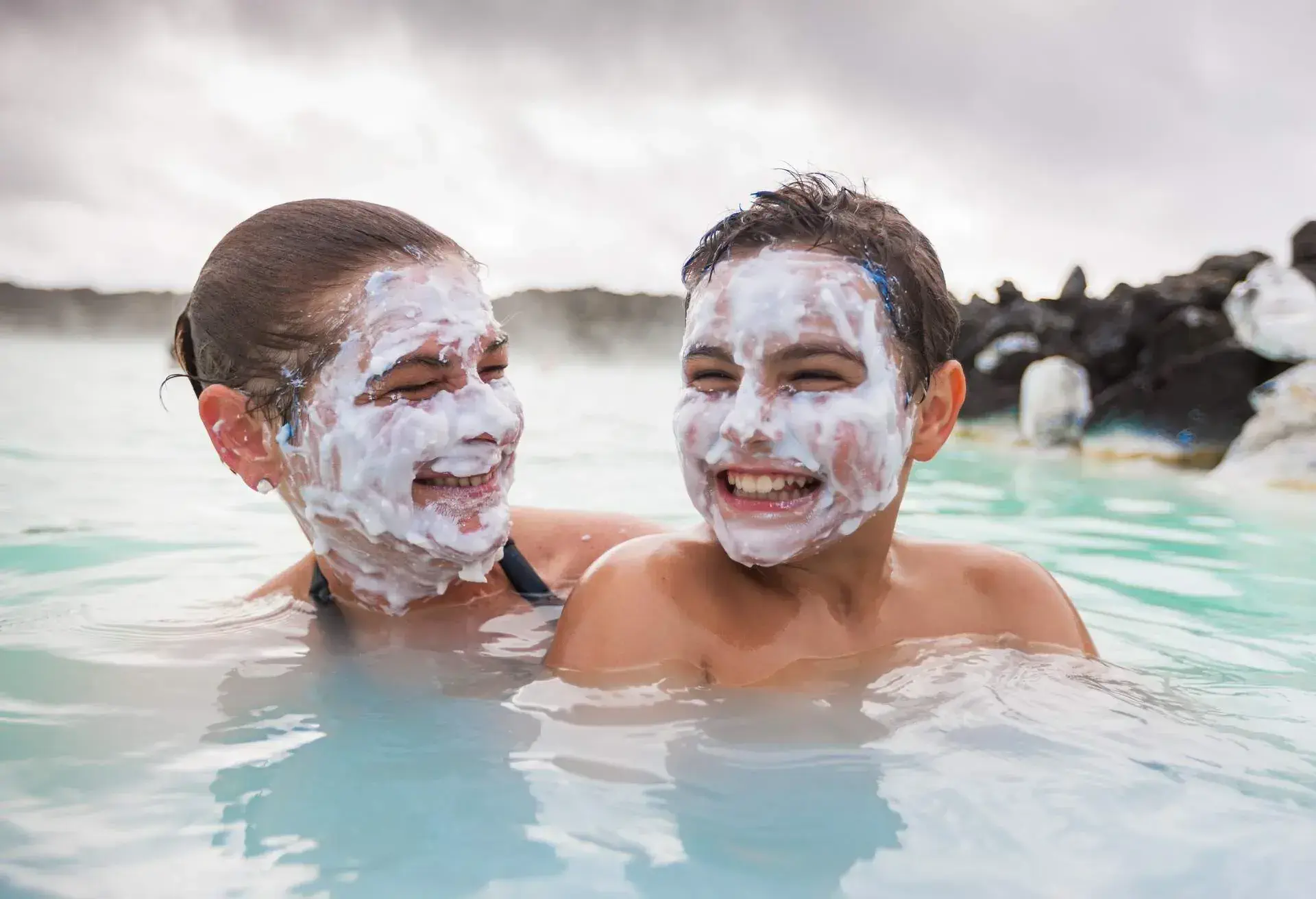 Mother and son having fun together, joking, smiling with natural sulphur face masks in Icelandic Natural Wellness Lagoon. Kevlavic, Iceland.