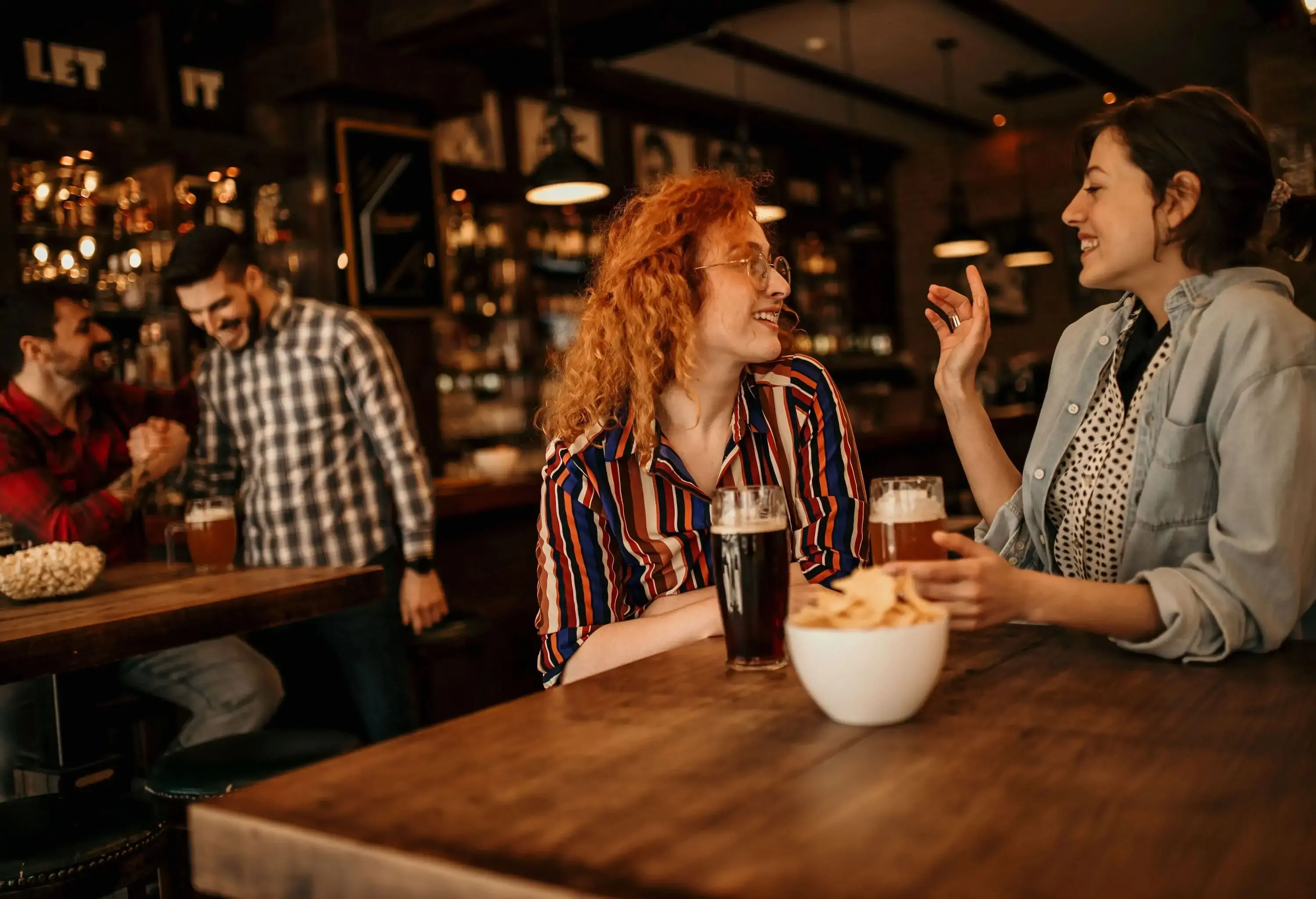 A pair of male and female friends sit at different tables with their snacks in a bar.