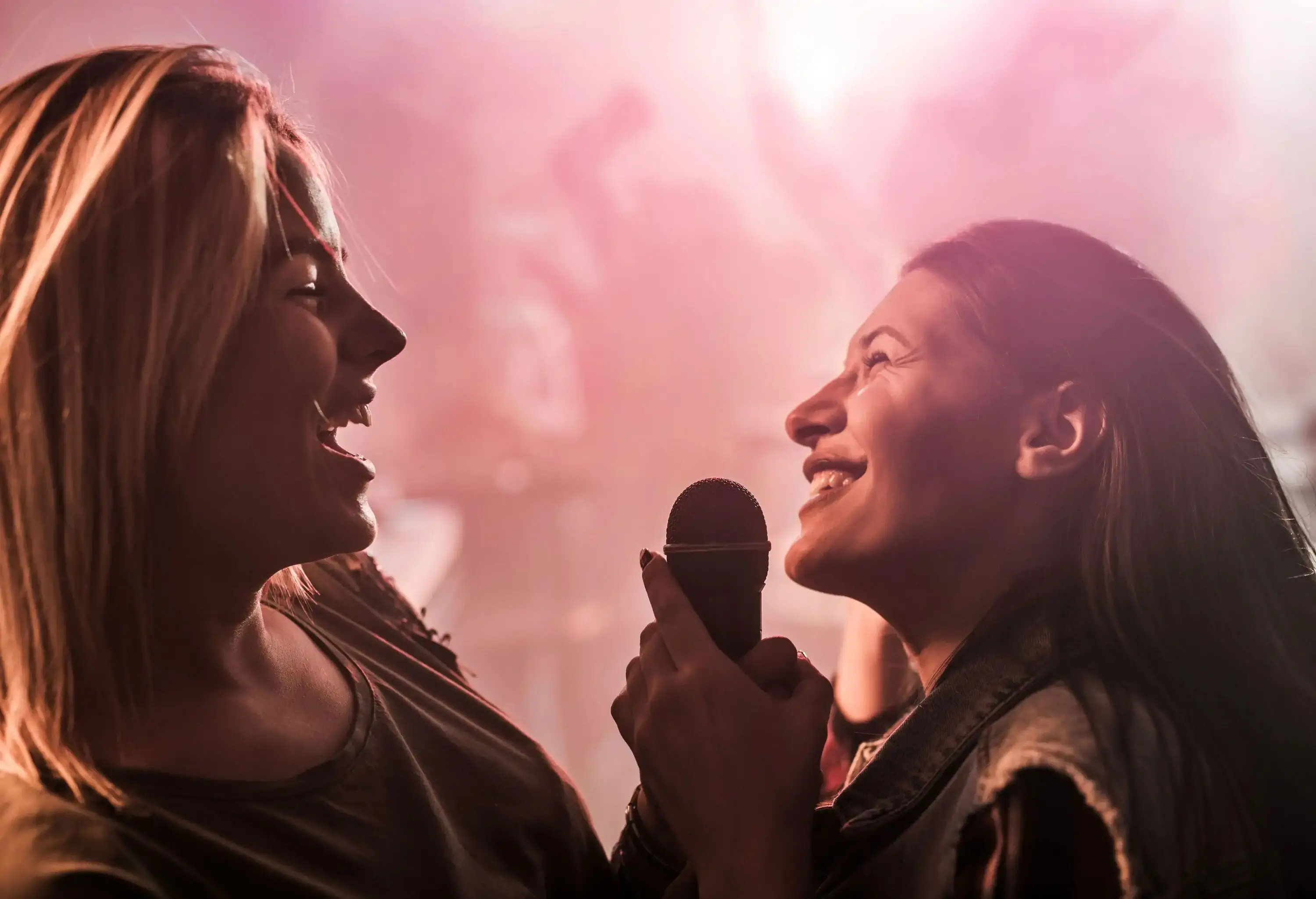 Two female friends happily sing into a single microphone in a bar.