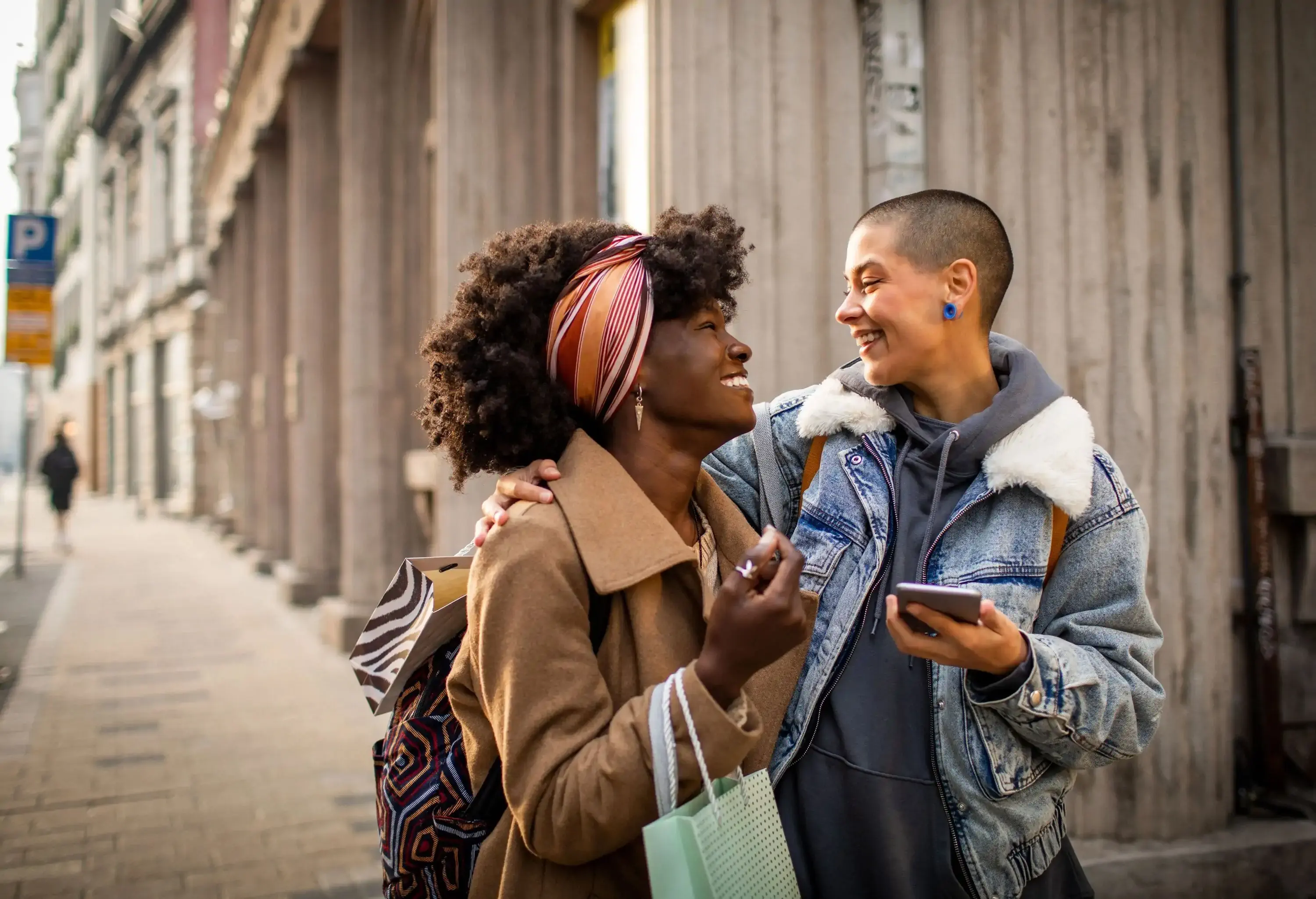 A curly-haired woman with a bandana and brown coat smiles at a woman with a crew cut hair and denim jacket.