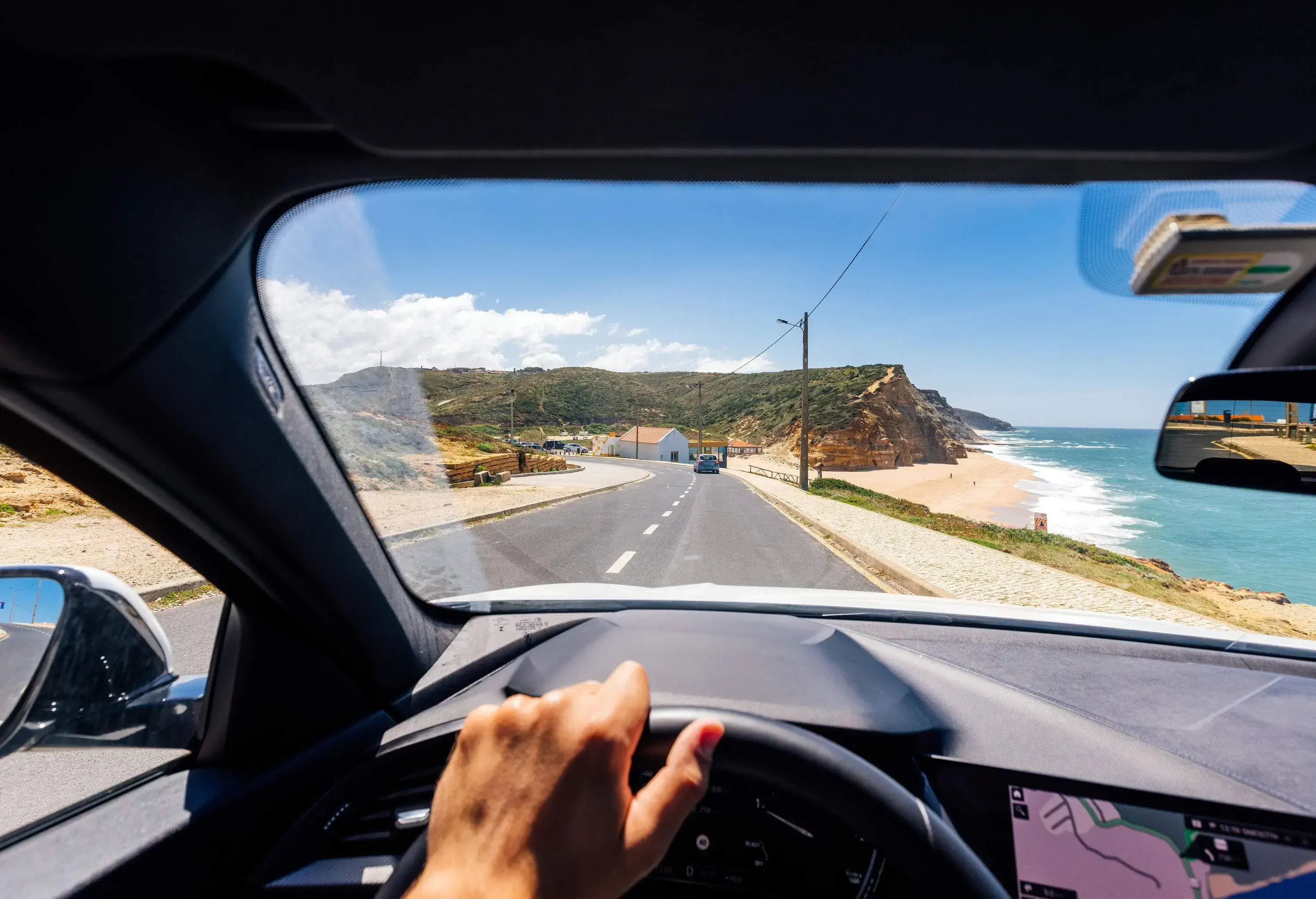 A hand on a car's steering wheel with a front view of a coastal road.
