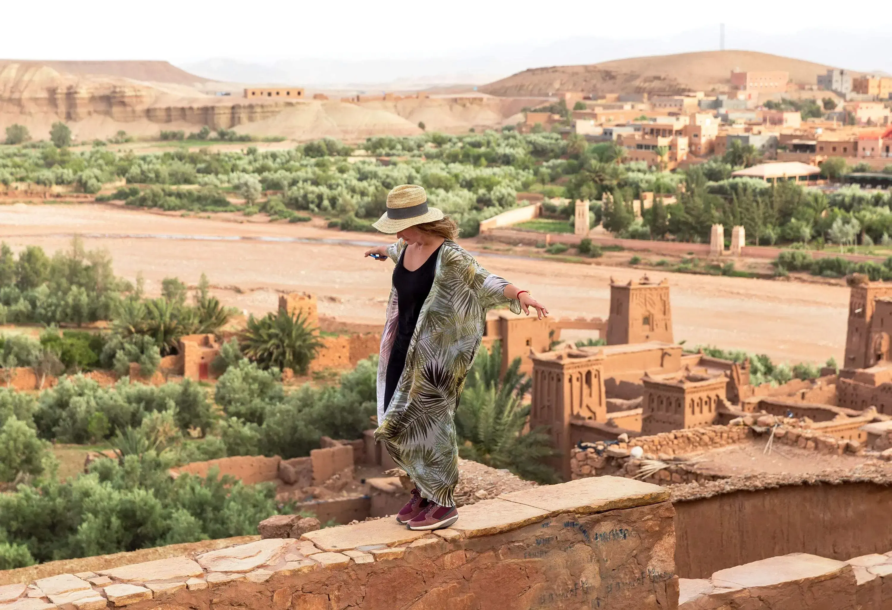 A girl with a brown hat balancing on the narrow edge of a wall with a view of traditional houses in the desert surrounded by green trees.
